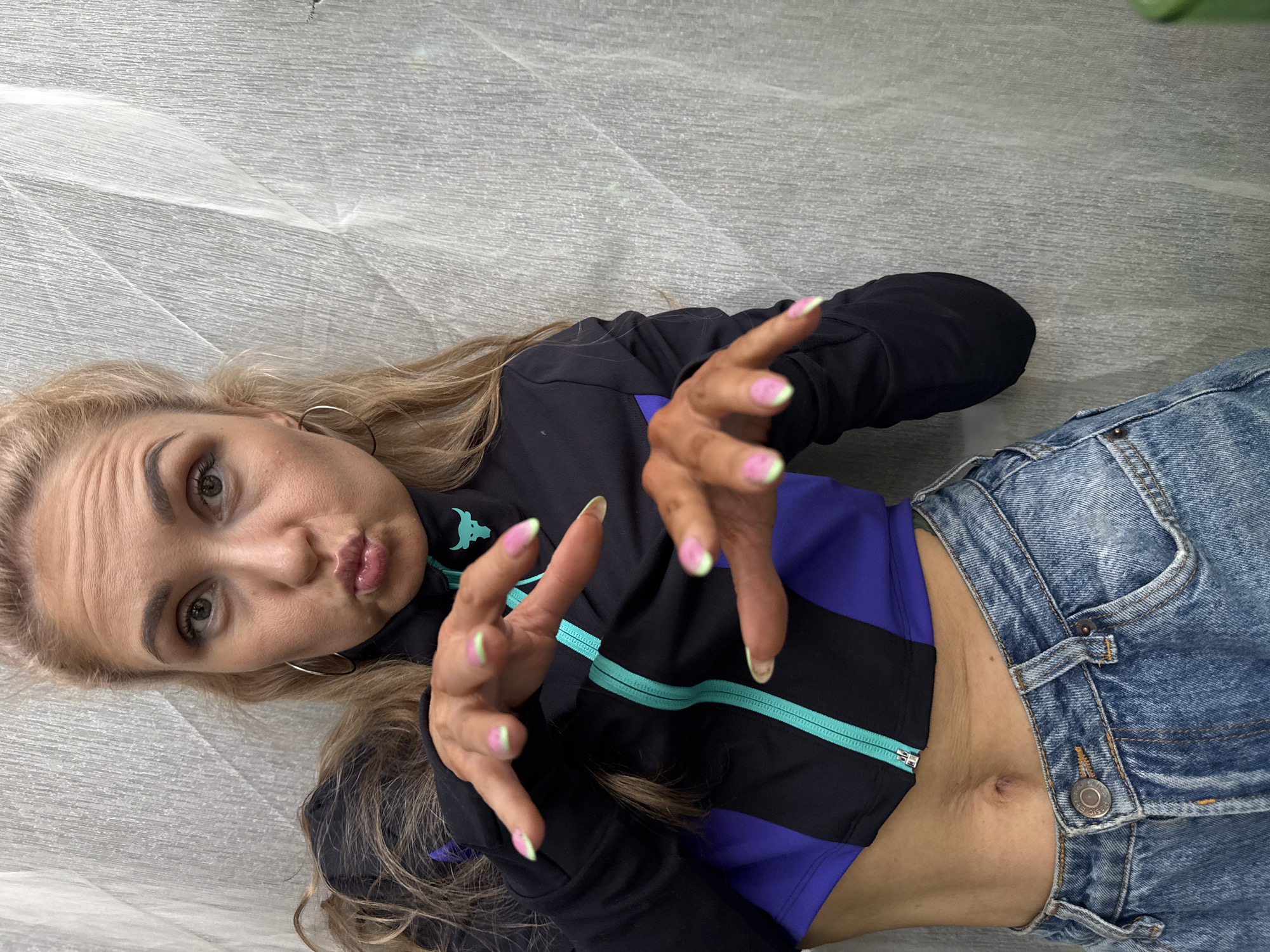 A young woman with blonde hair is depicted lying on a light gray, textured surface, possibly a floor or fabric. She is wearing a black and purple sports jacket with a turquoise zipper and light blue denim jeans, with her midriff exposed. Her hands are raised towards the camera, showcasing a unique manicure with pink polish and light green tips on her fingernails.
The woman is looking directly at the viewer with a playful, pouting "duck face" expression. Her eyes are open, and she has visible eyebrow makeup. She is also wearing silver hoop earrings. A small, stylized bull logo, resembling the Under Armour "The Rock" Brahma bull, is visible on the upper left chest area of her jacket. The image is taken from a slightly elevated perspective, looking down at the subject. The lighting appears even, suggesting an indoor setting. No specific location cues for Chaikovsky, Russia, are visible in the image itself.