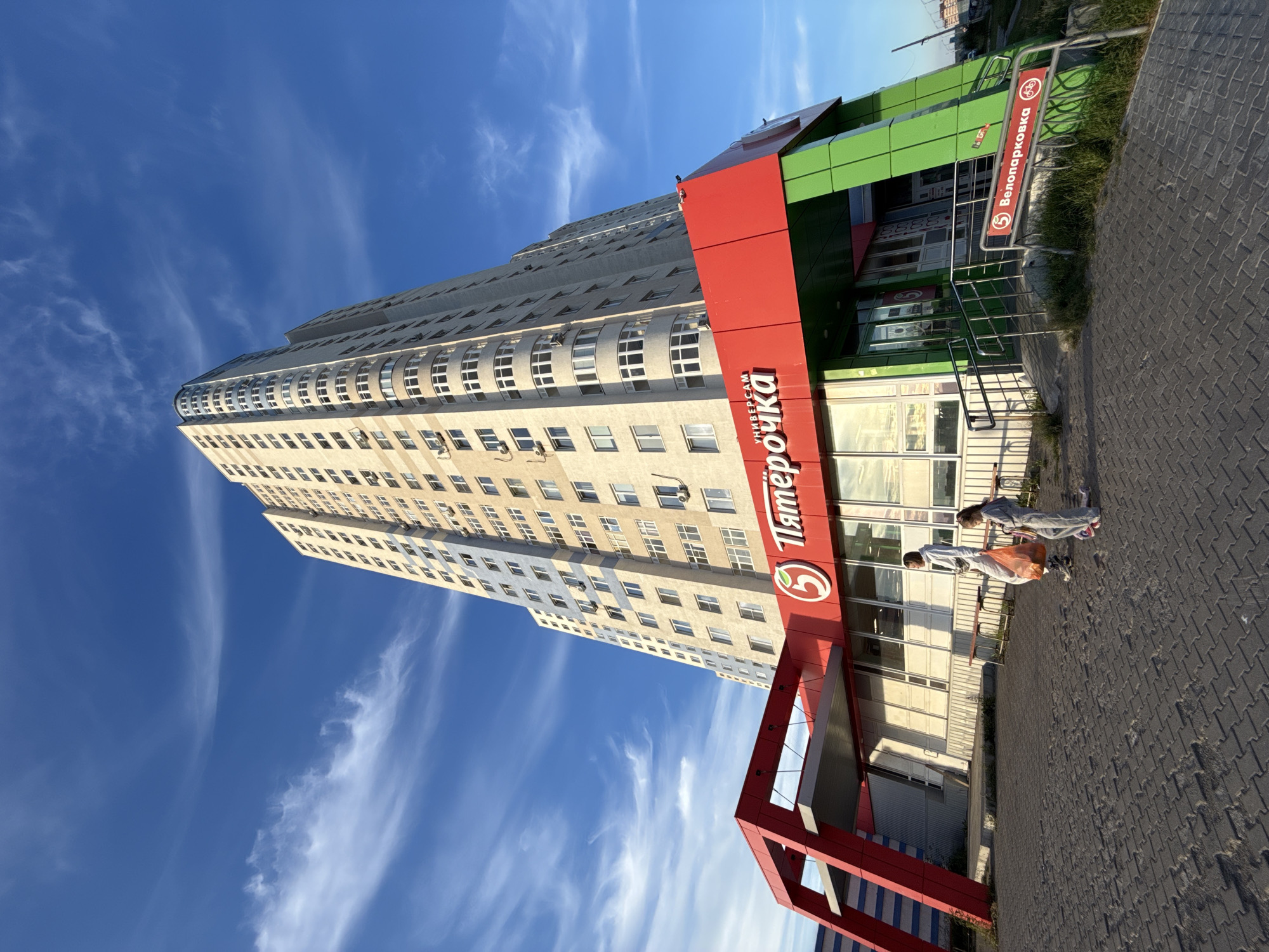 The image shows a tall, modern apartment building with many windows, under a bright blue sky with wispy clouds. In the foreground, a red and green facade of a store is visible, with a red sign displaying "Пятёрочка" (Pyaterochka), a Russian supermarket chain, and a bicycle parking sign in Russian that reads "Велопарковка" (Bicycle parking). Two individuals are walking past the store, one carrying a shopping bag. The sun appears to be setting or rising, casting long shadows. The scene is paved with interlocking bricks. The overall atmosphere is bright and urban.
