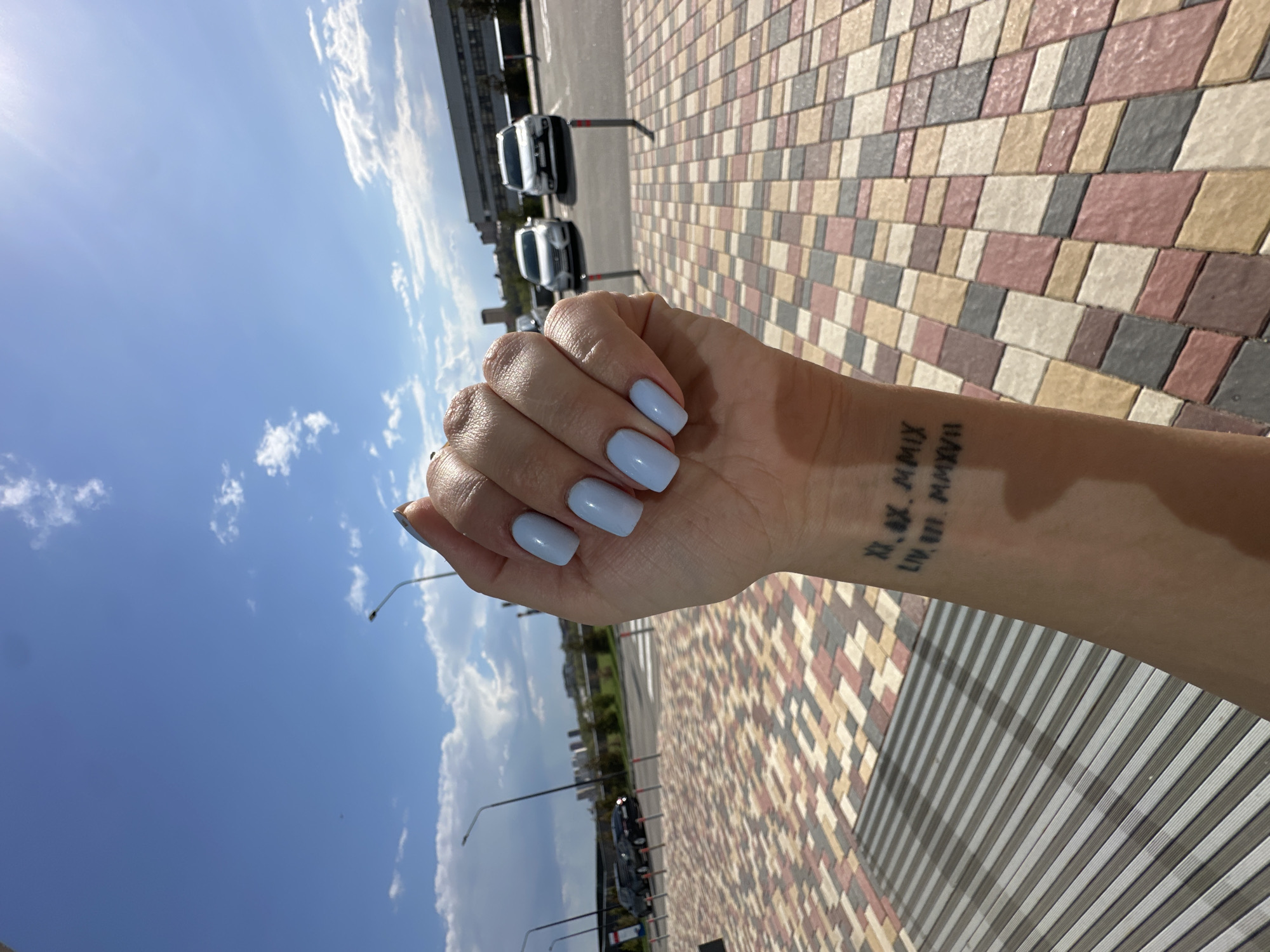 The image shows a person's hand and forearm against an outdoor urban background. The person has light blue nail polish. A tattoo is visible on the forearm. The background includes a patterned tile surface, a street with vehicles, streetlights, buildings, and a blue sky with clouds.