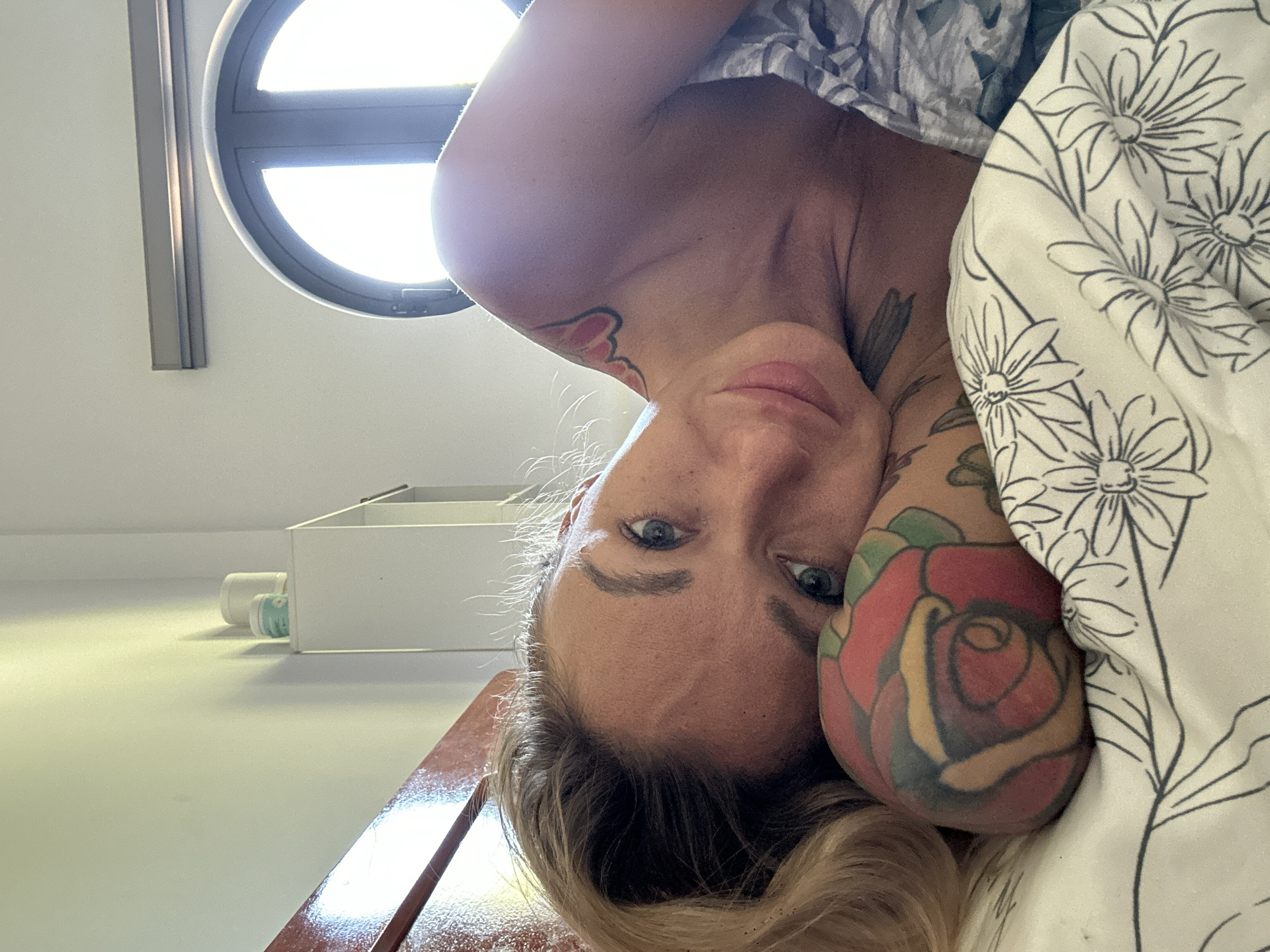 A blonde woman with blue eyes lies on a bed, propped up on her arm. She has tattoos on her arm and shoulder. The bedsheets have a floral pattern. In the background, there is a white cabinet with two containers on top, and a round window with sunlight streaming through. The scene appears to be indoors, possibly in a bedroom. The location in Mũi Né, Vietnam is not discernible from the image.