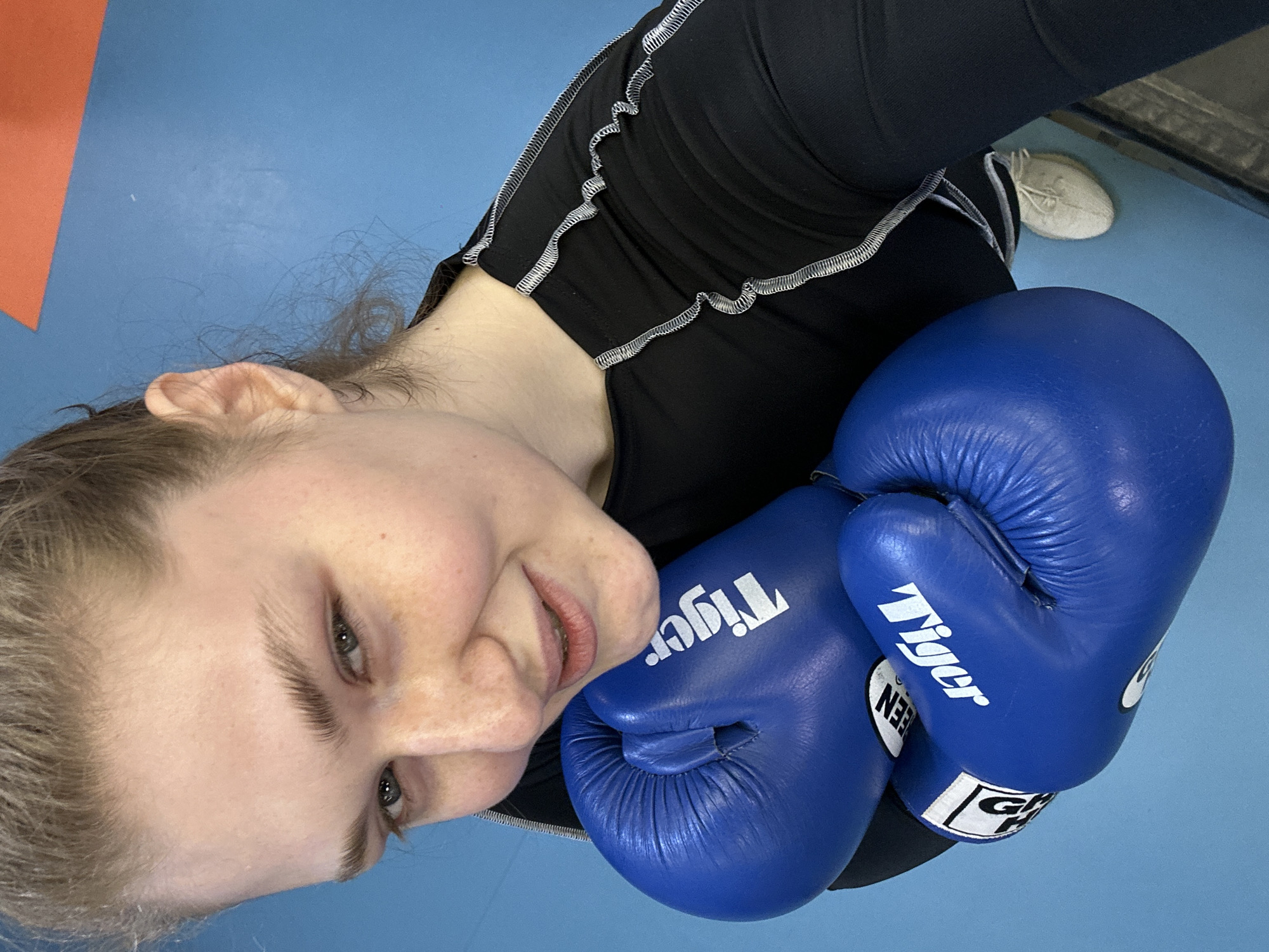 A young woman with fair skin and blonde hair holds up a pair of blue boxing gloves to the camera. She wears a black, long-sleeved athletic top with white stitching along the seams. A white tennis shoe is visible in the background. The setting appears to be a gym or fitness studio, as evidenced by the blue flooring and orange and grey wall sections. The brand names "Tiger" and "GREEN HILL" are visible on the boxing gloves. The specific location in Kazan, Russia, cannot be definitively determined from this image.