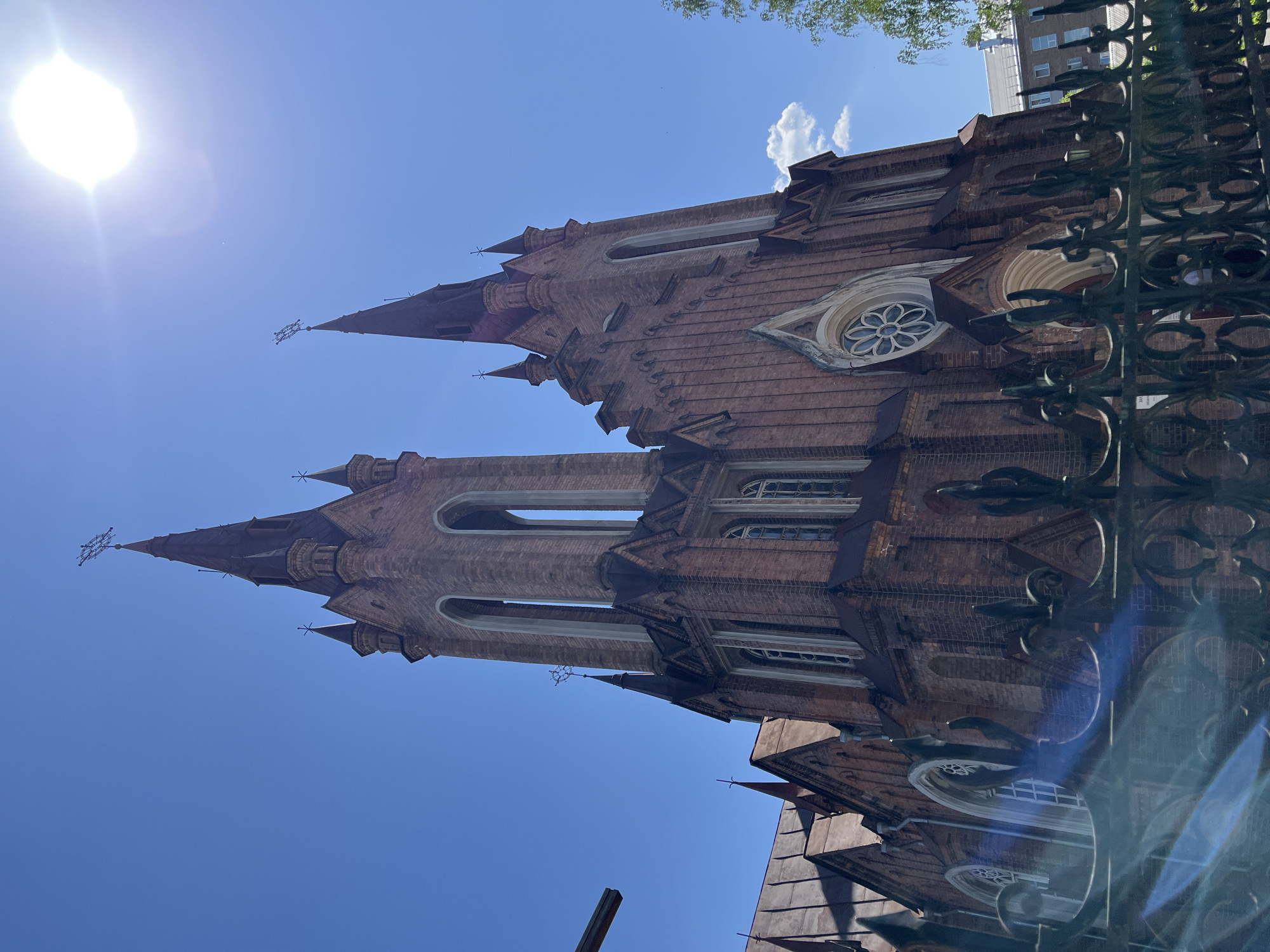 The image shows the Roman Catholic Church of the Transfiguration in Krasnoyarsk, Russia, viewed from a low angle, with the sun in the upper left corner. The church is constructed of red brick, featuring two prominent towers with pointed spires, each topped with a cross. Arched windows and decorative details are visible on the facades. A wrought-iron fence runs along the right edge of the frame, slightly blurred. The sky is clear blue with a few wispy clouds. A tall building and trees can be seen in the top right, behind the church.
