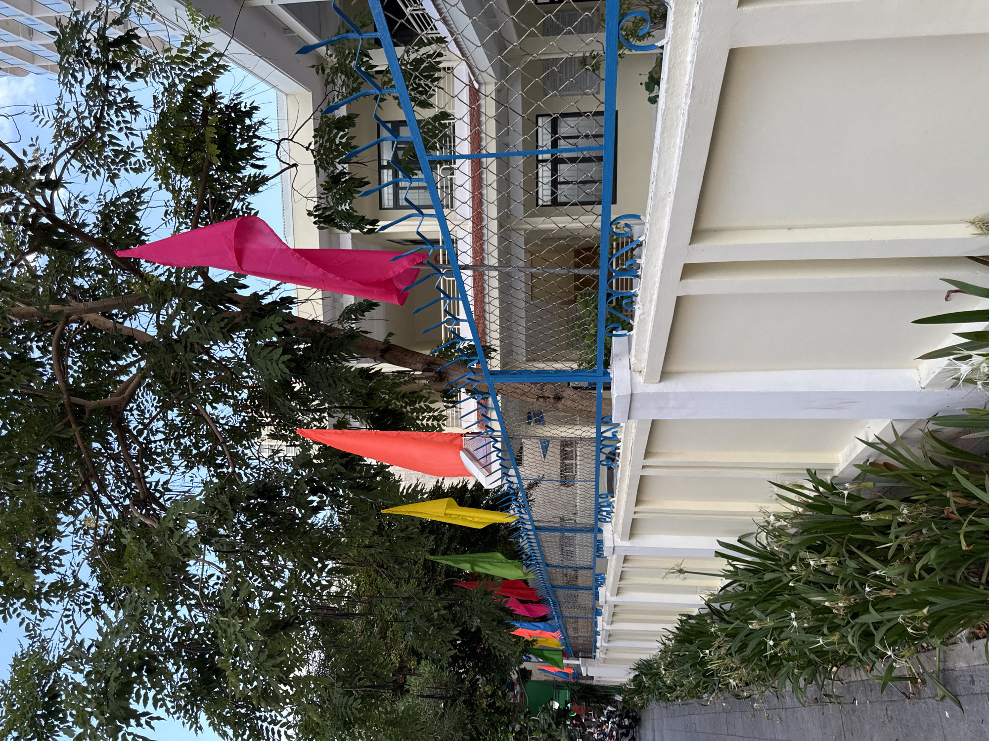 The image depicts a street scene in Nha Trang, Vietnam, featuring a row of colorful flags strung along a blue metal fence. The flags, in shades of pink, orange, yellow, green, and blue, are partially obscured by the lush green foliage of a large tree overhead. The fence itself is decorated with decorative finials. Behind the fence, a light-colored building with multiple windows and balconies is visible. The foreground shows a sidewalk with a line of green plants and white flowers running along the base of a cream-colored wall that forms the boundary of the property. A few motorcycles are parked on the left side of the sidewalk, suggesting an urban setting. The sky above is a clear blue.