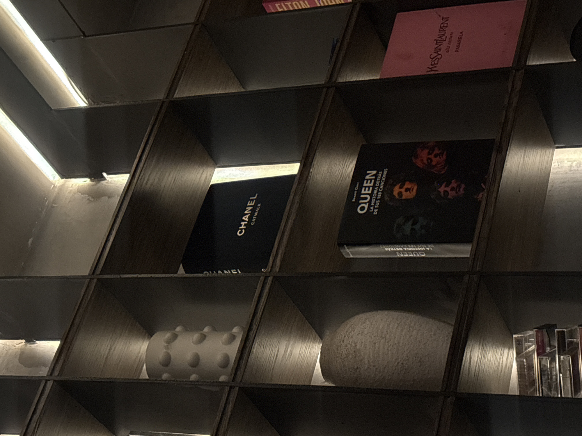 The image displays a detailed view of a modern, dark-stained wooden shelving unit featuring multiple square and rectangular compartments. Integrated strip lighting illuminates various sections from above, creating highlights and shadows.
In the upper-left compartment, a white, cylindrical decorative vase with evenly spaced circular protrusions is visible. Directly below it, in the mid-left compartment, an off-white, textured, ovoid vase rests.
The top-center shelf holds a black book titled "CHANEL CATWALK" in white lettering. To its right, partially obscured, is a book with a red or orange spine displaying "ELTON JOHN."
The central-right compartment showcases a black book titled "QUEEN LA HISTORIA DETRÁS DE SUS 188 CANCIONES" by Benoît Clerc, featuring a cover image of the four Queen band members. Adjacent to it, on the far right, is a prominent pink book titled "YVES SAINT LAURENT" with additional text "alta costura" and "PASARELA" in black script.
The bottom-left visible compartment contains a stack of several clear plastic cases, likely holding cassette tapes or similar media.
The overall environment, characterized by curated luxury brand books (Chanel, Yves Saint Laurent) and iconic music biographies (Queen, Elton John), along with sophisticated integrated lighting, suggests a high-end interior. The Spanish text on the "QUEEN" and "YVES SAINT LAURENT" books indicates a Spanish-speaking locale. This setting is consistent with a luxury hotel lobby, lounge, library, or an upscale boutique in Mexico City, Mexico, likely in areas such as Polanco, Roma Norte, or along Paseo de la Reforma.