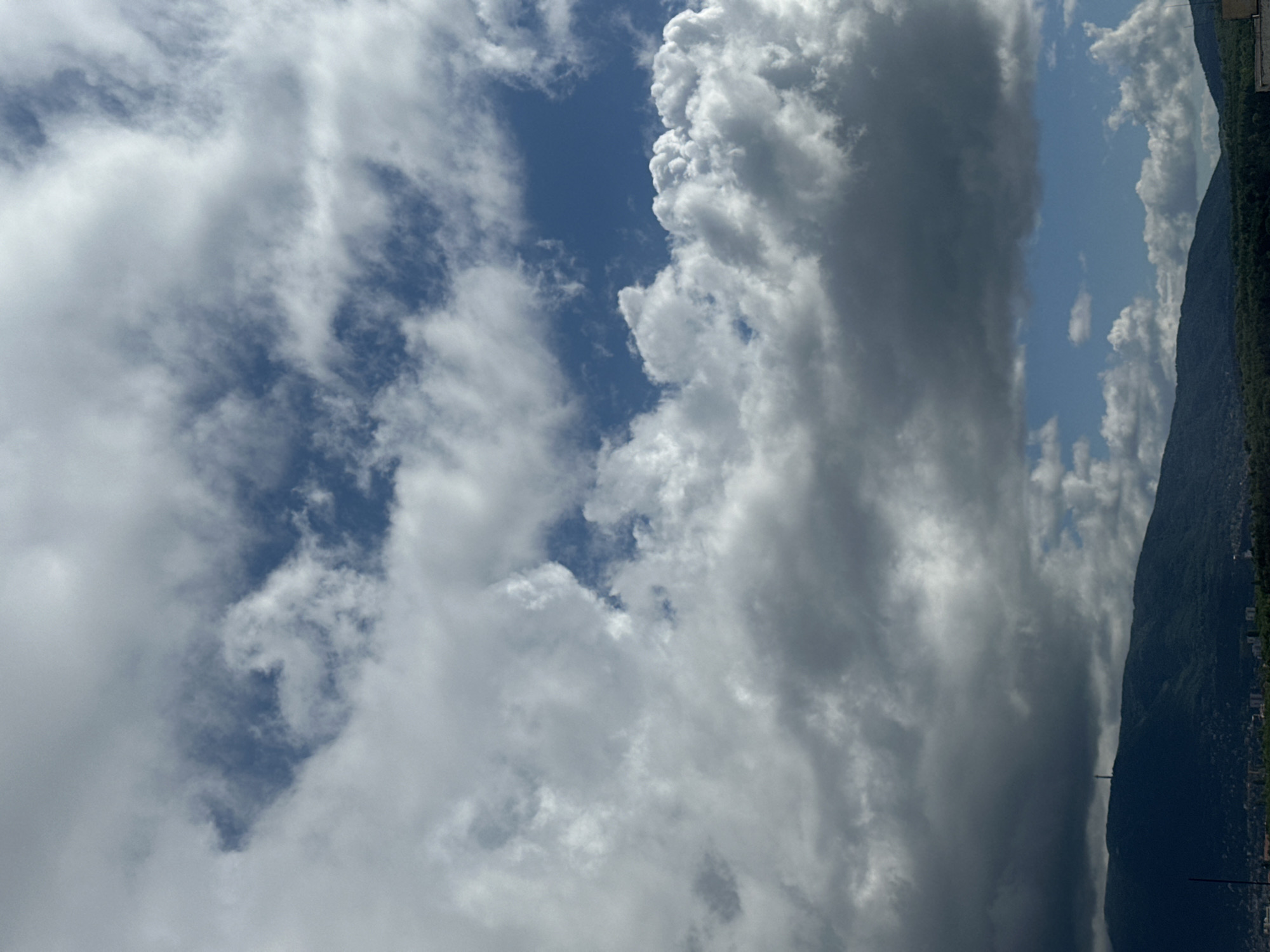 The image captures a dramatic sky over a mountainous landscape during daytime. The scene is dominated by a diverse cloudscape, with a large mass of dark, heavy grey clouds occupying the right side of the frame, suggesting dense cover or potential weather. To their left, bright white, fluffy cumulus clouds are interspersed with clear, vibrant patches of blue sky, creating a strong contrast. Lighter, wispy clouds are also scattered across the upper left.
Below the sky, a dark green, heavily forested mountain range stretches across the bottom of the image, forming the horizon. Faint outlines of urban development or buildings are visible along the base of the mountain, indicating a city or town. A distinct, slender tower-like structure stands prominently on the mountain's slope. The bright sky and sunlit clouds suggest it is late morning or afternoon. The mix of clear and stormy-looking clouds indicates variable weather. There is no discernible activity or visible text in the image.
Given the location context, the mountain is likely Vitosha Mountain, overlooking the city of Sofia, Bulgaria, and the tower could be the Kopitoto TV Tower.