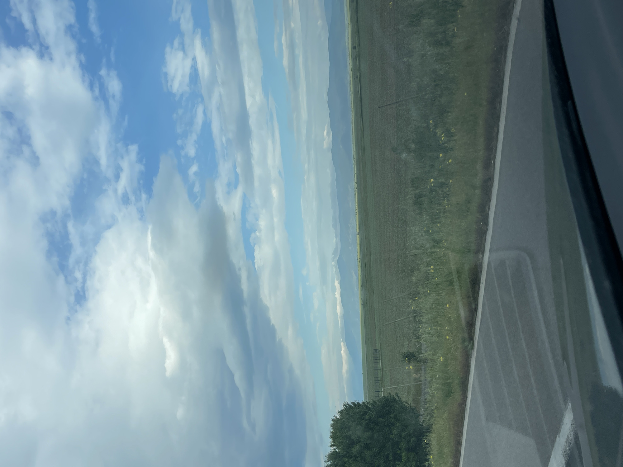The image depicts a view from a vehicle, likely a car, driving along a road near Vodenichane, Bulgaria. The sky is predominantly blue with layered white clouds. A field extends to the right, punctuated by distant fence posts and hints of yellow wildflowers. Beyond the field are indistinct mountains. A large green tree is visible in the lower part of the frame. Part of the vehicle's interior and window is visible on the right edge. There are no people visible in the image.
