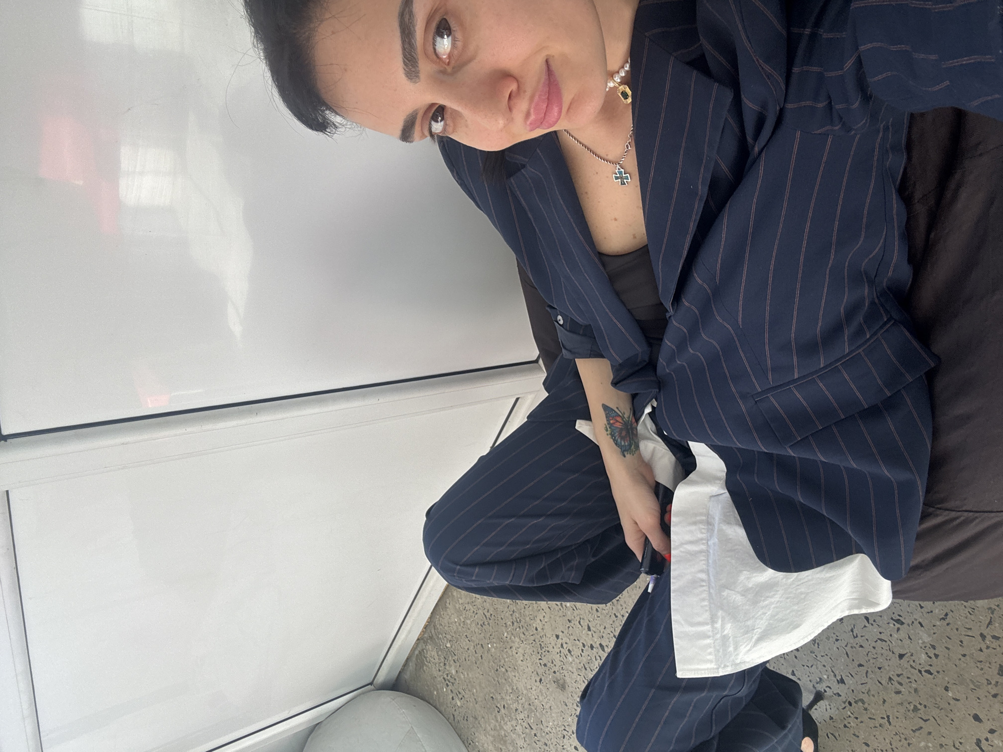 A medium-shot, slightly tilted portrait features a woman posing indoors in Tbilisi, Georgia. She looks directly at the camera with a subtle, perhaps enigmatic, expression on her face, featuring a slight pout or smirk. Her dark hair is pulled back, and she has prominent dark eyebrows.
The woman is wearing a navy blue pinstriped suit, consisting of a blazer and matching trousers. A dark top is visible underneath her blazer, and a white garment, possibly a shirt or part of the blazer's design, is seen near her left hand. She accessorizes with two necklaces: one made of pearls with a gold-colored charm, and another silver-toned chain featuring a cross pendant with a blue or green inlay. A vibrant, multi-colored butterfly tattoo is visible on her left forearm/wrist. She holds a dark, pen-like object in her left hand.
She appears to be seated on a dark brown, upholstered surface, possibly a beanbag or cushion, which occupies the right side of the frame. The floor is a light gray, speckled material, suggesting concrete or terrazzo. To her left, a bright white wall or partition features faint reflections of what seem to be internal window frames or structures, indicating an indoor setting with good natural light during the daytime. No visible text is present in the image.