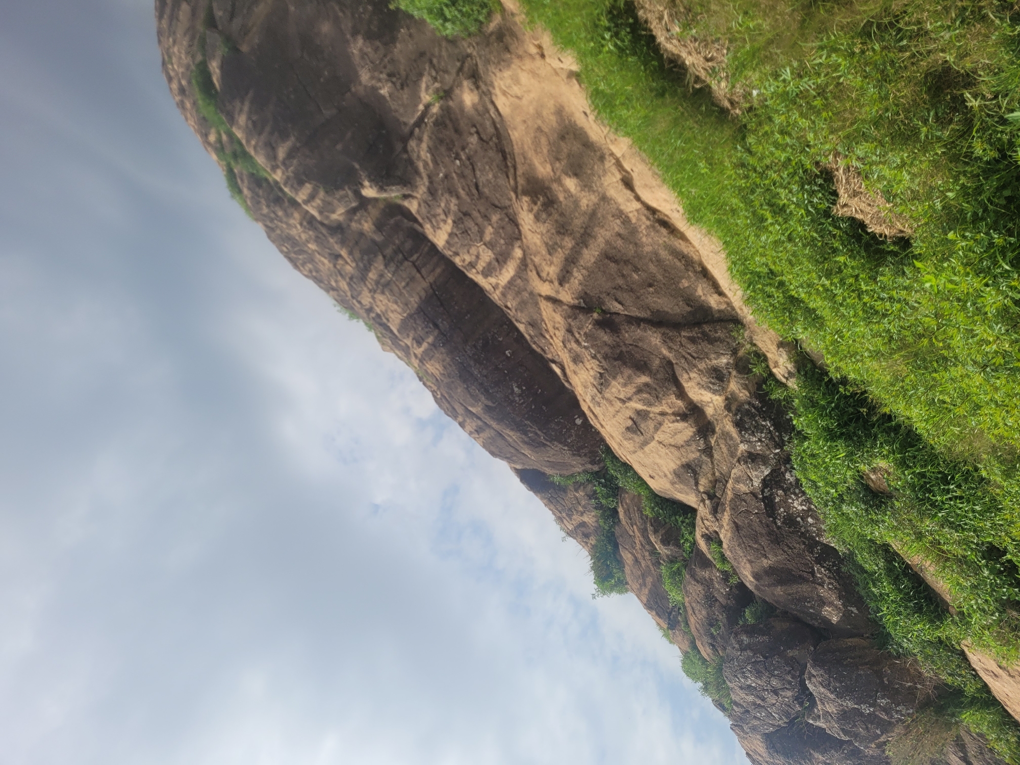 A low-angle, outdoor, full shot captures a rocky hillside with patches of vibrant green grass and some foliage. The rocks vary in color from light tan to dark brown, exhibiting a rough texture and visible stratification. The sky above is overcast with light grey clouds. The scene is located in Bukuru, Nigeria.
