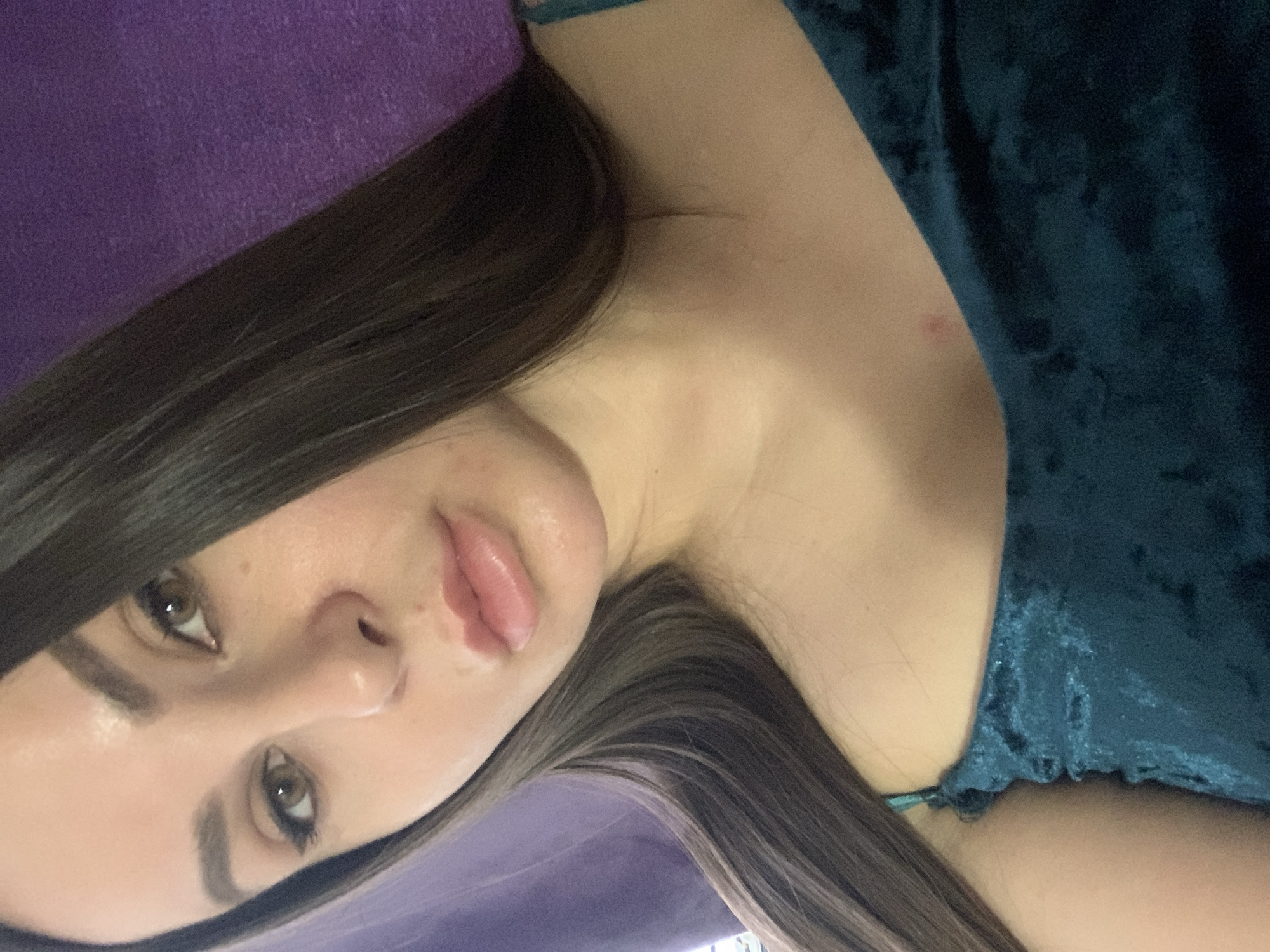 A close-up, waist-up, indoor, selfie-style photograph shows a young woman with dark hair and light brown eyes. She is looking directly at the camera with a neutral expression. She is wearing a dark teal, velvet, strappy top. The background is a blurred, textured purple wall. The lighting is soft and even, with a slight glow on her skin.