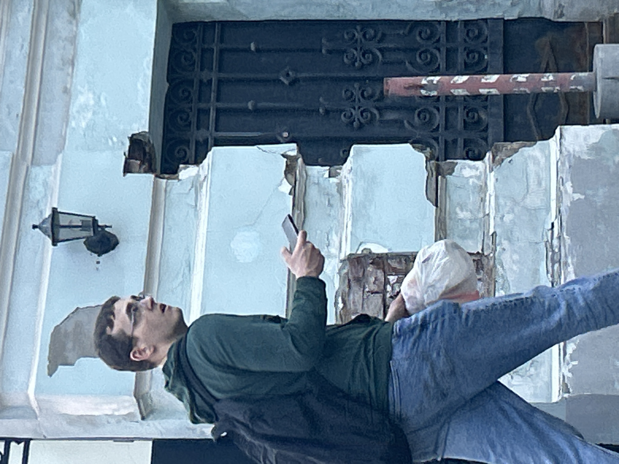 A man is shown in the foreground, standing and looking up while holding a cell phone in his right hand. He's wearing blue jeans, a dark green long-sleeved shirt, and appears to have a dark-colored bag slung over his shoulder. Behind him is a building with significant signs of disrepair, including cracked and crumbling surfaces. The building has a wrought-iron gate and a lamp fixture attached to the wall. The setting appears to be in a city, but the specific location is not identifiable from the image.