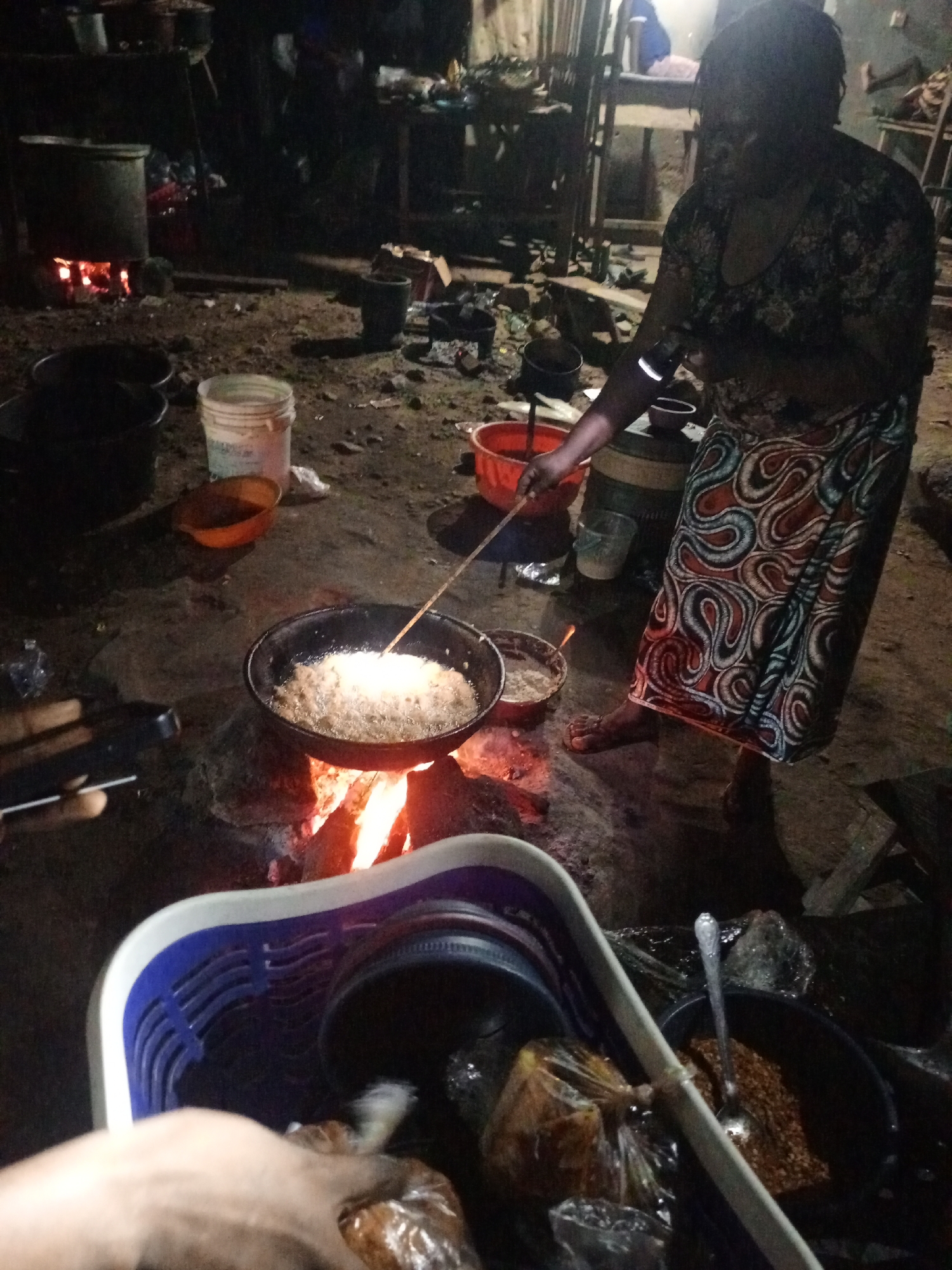 In a dimly lit outdoor space, possibly in Keffi, Nigeria, a woman is cooking over an open fire. She is standing barefoot, wearing a patterned top and a long, colorful skirt. She stirs food in a large frying pan over the fire with a long stick. To her left, a red bowl sits on a barrel, while another smaller bowl sits on the ground. A larger pot is on a separate fire in the background. In the foreground, a blue and white basket contains various wrapped items and a pot with a spoon inside, indicating food preparation. Another individual is seen holding what may be a phone near the bottom left corner of the image. The environment seems to be an open-air cooking area, possibly a roadside food stall.