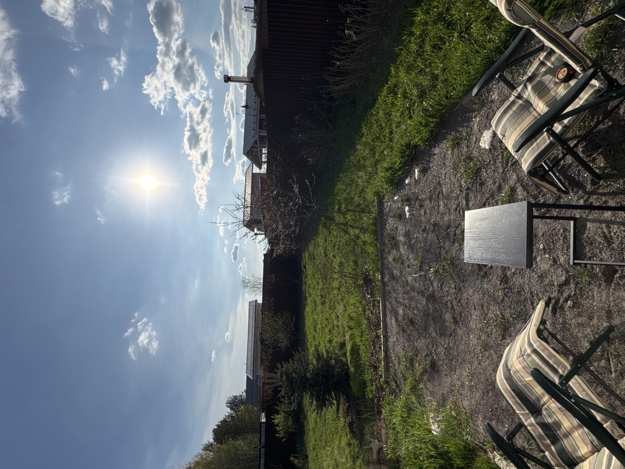The image depicts an outdoor scene in Steklyannyi, Russia. Two striped lawn chairs surround a small, dark grey, square table. The ground is a mixture of dirt and gravel. A fence runs along the edge of a green grassy area. In the background, partially obscured by the fence, is a multi-story house. The sky is blue with scattered white clouds, and the sun is brightly shining. There are no people visible in the scene.