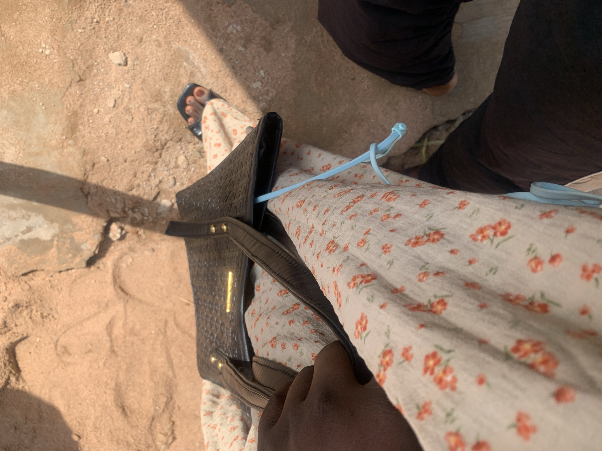 The image captures a close-up, overhead view of a person's legs and torso, seated outdoors on sandy ground. The person is wearing a cream-colored dress with a small red floral pattern and dark pants. A black handbag with gold hardware is placed on their lap. A light blue cord or strap is visible hanging from the handbag. The ground is composed of dry, sandy earth with some small stones. The lighting suggests a sunny day, casting shadows on the ground. The scene appears to be a simple outdoor seating area, potentially a roadside or waiting area, in Jalingo, Nigeria.
