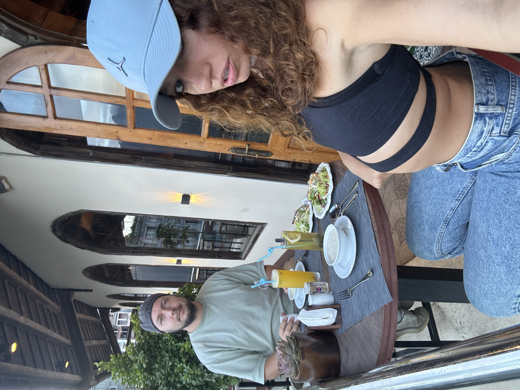 Two individuals are at a table on a balcony, presumed in Sa Pa Ward, Vietnam, given the architectural features in the background. The woman, wearing a black top, light-wash jeans, and a light blue baseball cap, is holding a phone or camera, presumably taking a photo. She has curly hair. The man, wearing a grey cap and a light green shirt, is seated at the table. The table is set with two drinks, two salads, soup, napkins, cutlery, and a potted succulent. The backdrop includes windows, a building, and some greenery.
