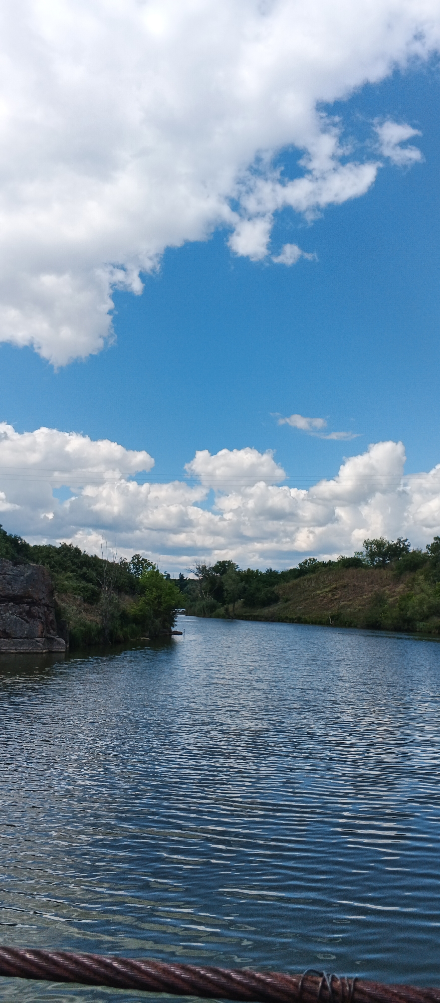 The image depicts a scenic view of a river or canal under a bright, cloudy sky. On the left bank, there are large rocks and dense green foliage. The right bank features a gentle slope covered in dry grass and scattered trees. The water's surface is rippled, reflecting the blue sky and white clouds. In the foreground, a thick, rusted cable is visible, suggesting it is part of a bridge, pier, or some form of industrial structure. The scene is calm and natural, with a hint of human infrastructure. Based on the general landscape and vegetation, this location could be along the Ternivka River in Ternivka, Ukraine, a region known for its industrial history and natural waterways.