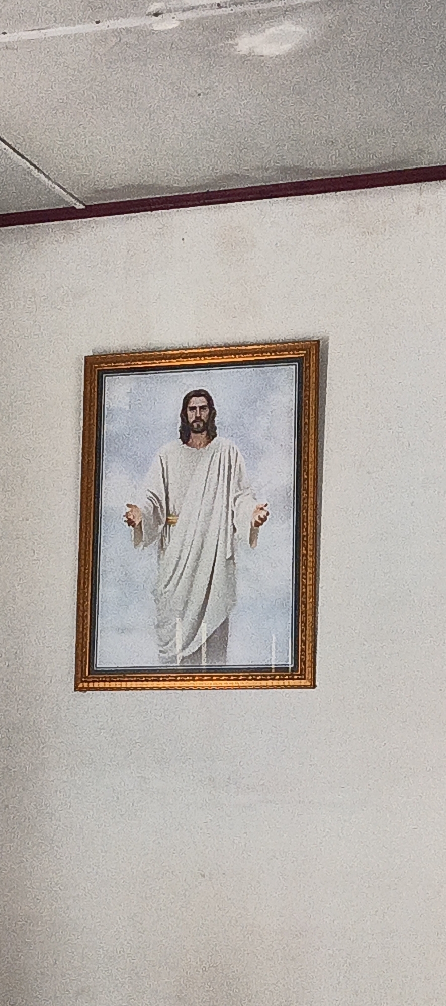 A framed painting of Jesus Christ hangs on a textured, light-colored wall. The painting depicts Jesus from the waist up, with long, dark hair and a beard. He is wearing a white robe draped over his body and has his arms slightly extended, palms facing upwards. The background of the painting is a light blue, suggesting a sky or ethereal setting. The frame is ornate, with a golden hue. The wall above the painting appears to have a ceiling or upper wall section with some discoloration and a dark line running horizontally. This scene is located in the city of Cinta Dame, Indonesia.
