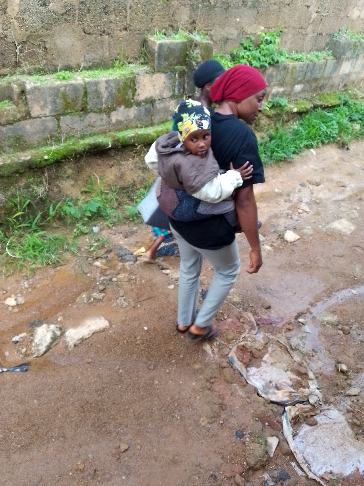 A woman wearing a black t-shirt and a red headscarf carries a baby on her back. The baby is wearing a brown jacket and a colorful hat. Another person wearing a black headscarf is partially visible behind the woman. They are walking on a dirt path with some puddles and debris. In the background, there is a stone wall with some vegetation growing on it. The scene appears to be in an outdoor, possibly rural or less developed, environment.