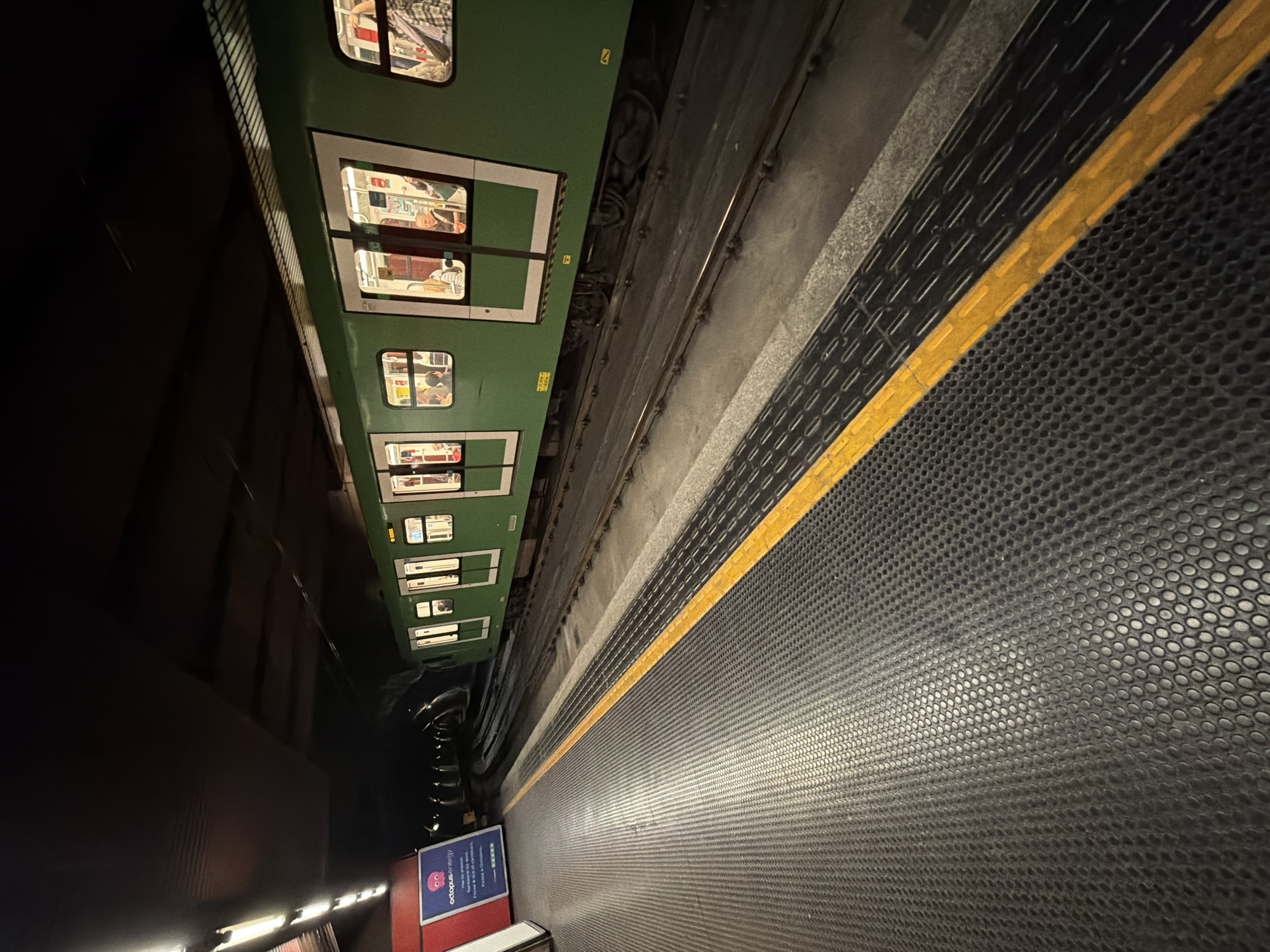 The image shows the Milan Metro (subway) platform. A green train is at the station. Passengers are visible inside the train cars. The platform is dark and covered in a rubber mat. A yellow safety line runs along the edge of the platform. A sign, in Italian, is visible on the platform. The photo appears to be taken at night.