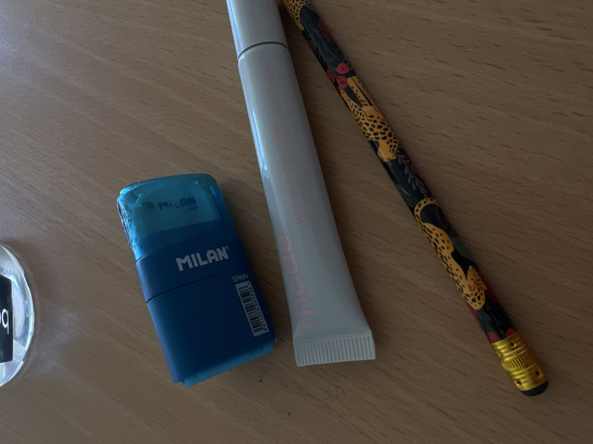 The image shows a wooden surface with several objects. There is a blue pencil sharpener with the word "MILAN" printed on it and the word "SPAIN" written next to a barcode. A gray tube of Rhode Peptide Lip Treatment is beside the sharpener. A black pencil with a leopard print and a gold eraser cap is to the right of the tube. A blurry glass object with the letter 'b' is visible on the left side. The scene does not suggest any information about Budva, Montenegro.