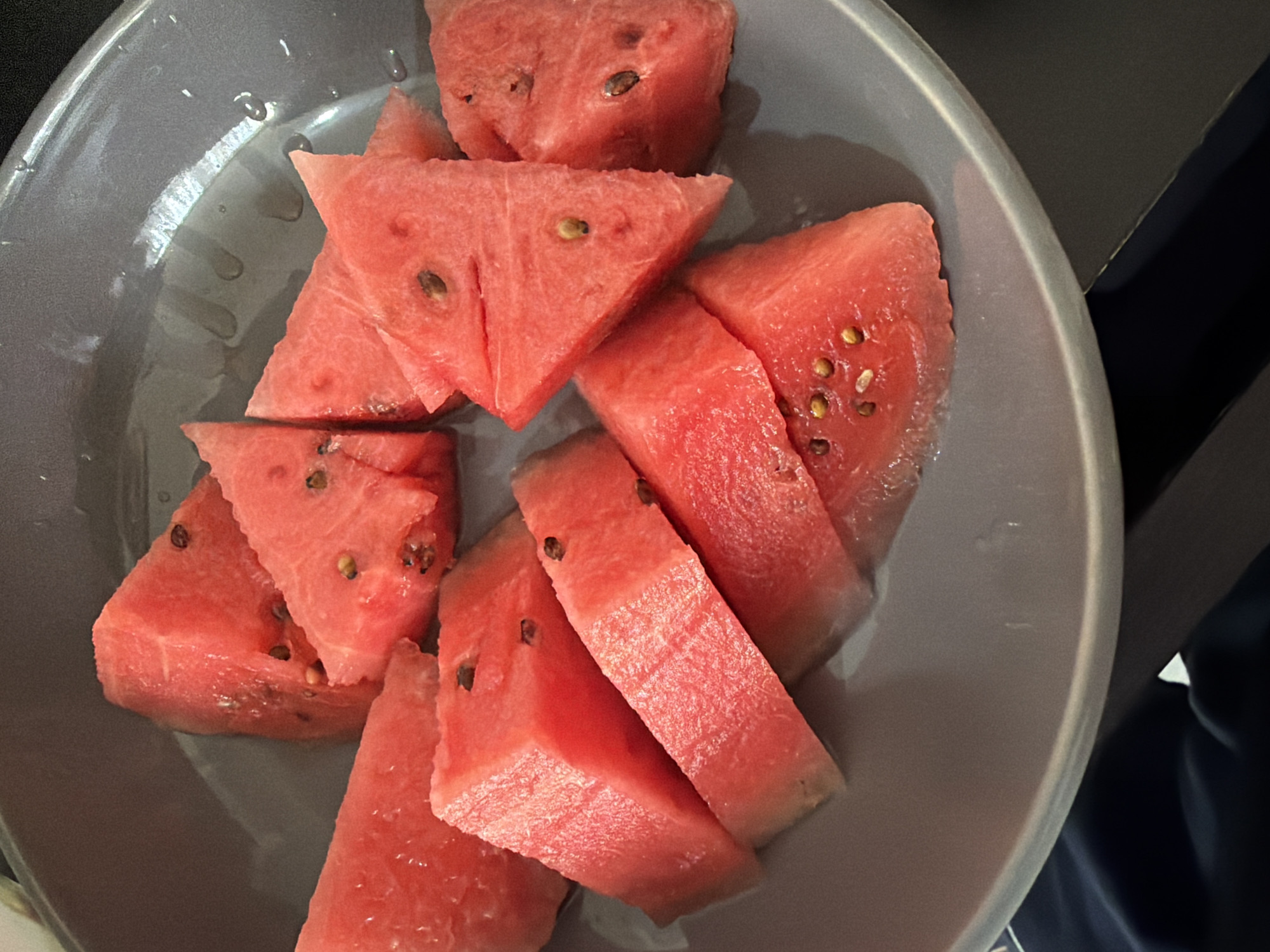 A top-down close-up shot depicts a collection of irregularly cut pieces of watermelon, exhibiting a vibrant pink hue, arranged haphazardly within a grey bowl. The fruit pieces display varying amounts of black seeds embedded in the flesh. Condensation or moisture is visible on the surface of both the watermelon and the bowl. The bowl itself is situated on or near a dark surface, likely a table or countertop. The scene is indoors. There is no evidence to suggest this image was taken in Pentre, United Kingdom.