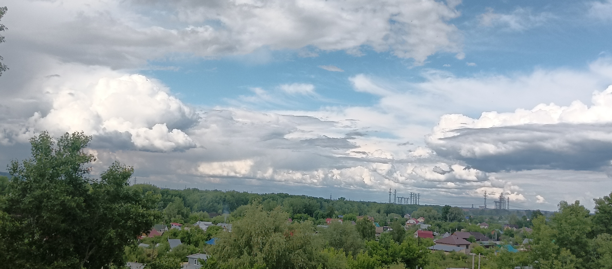 The image depicts a landscape under a cloudy sky with scattered blue patches. In the foreground, lush green trees frame the view, suggesting an elevated perspective. Below the trees, a residential area is visible, with houses nestled among the foliage. In the distance, power lines and industrial structures stand against the horizon. The location appears to be a suburban or rural area with industrial activity in the vicinity.