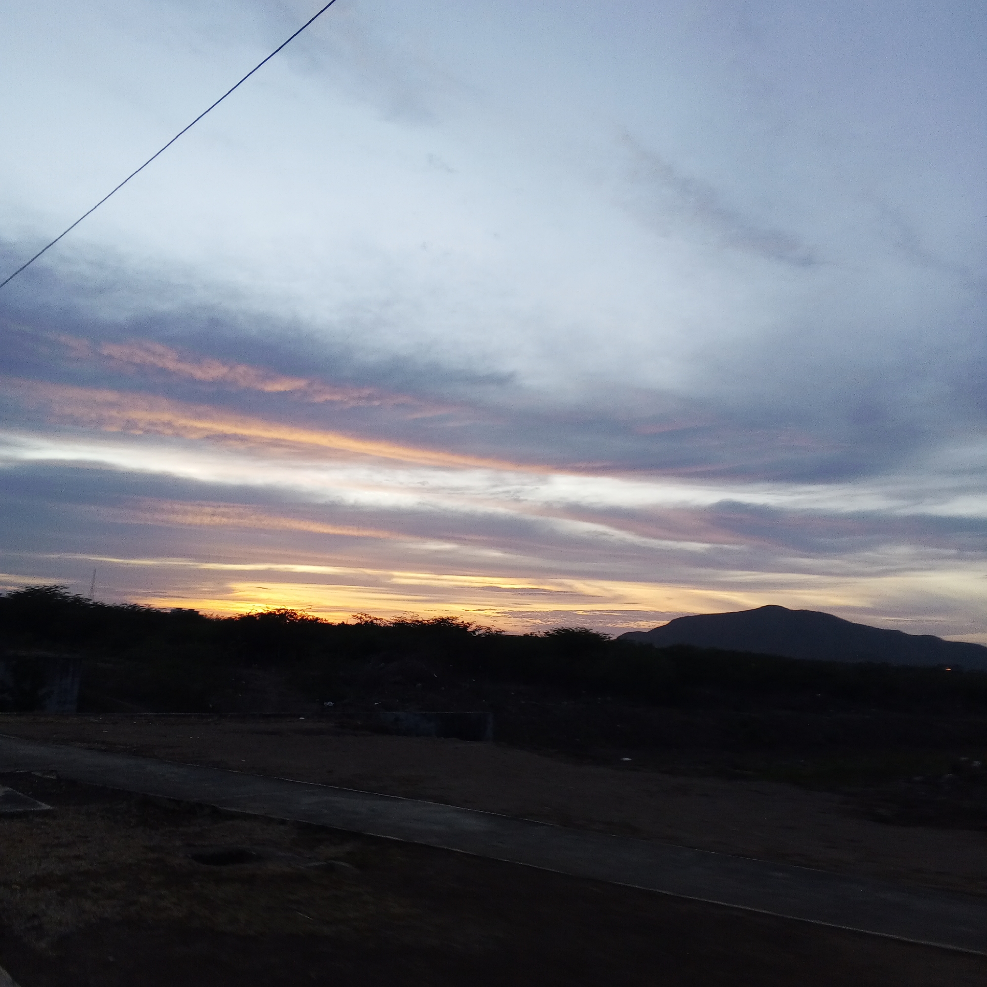 The image shows a landscape at dusk, possibly near the city of Unknown, Unknown. In the foreground, there is a road and some dark, undefined terrain. A line of trees or bushes is situated between the road and the horizon. Beyond this vegetation, a mountain is visible. The sky is filled with clouds, with a gradient of colors indicating the sunset or sunrise. The clouds range from light grays and whites to shades of orange and pink near the horizon. A single cable stretches across the upper left of the frame.