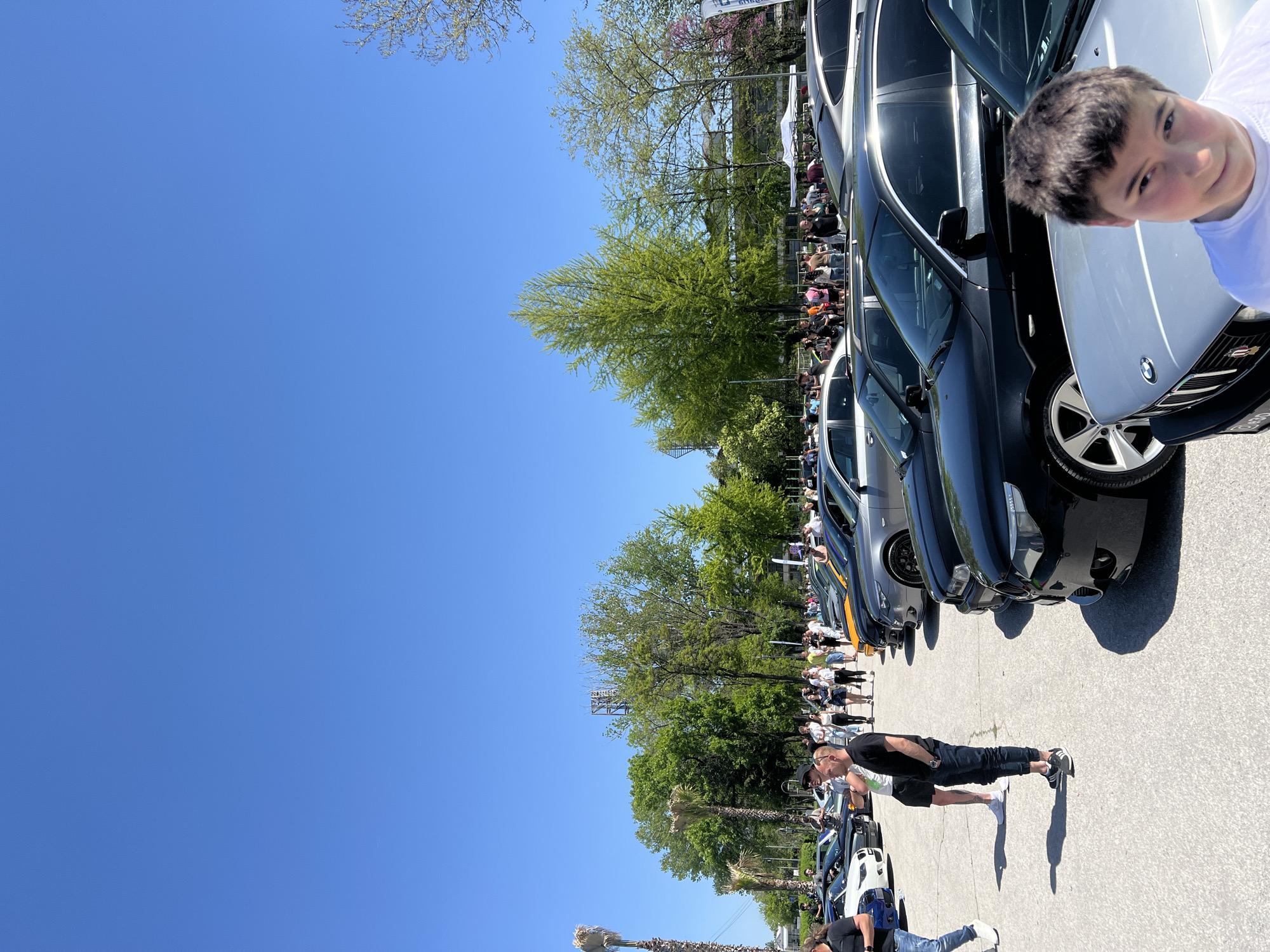 The image shows an outdoor event in Plovdiv, Bulgaria, featuring a car show or gathering along a paved area. Numerous people are visible, mostly standing or walking near the cars. A variety of parked cars, mostly modern models and mostly dark colors, line the right side of the frame. Some people are close to the cars, interacting and looking at them. A boy is close to the camera, near a white car. Behind the cars, trees and some green foliage line the area, providing a natural backdrop. A clear, blue sky fills the upper left portion of the image. The location appears to be an organized car event in an open-air space within the city.