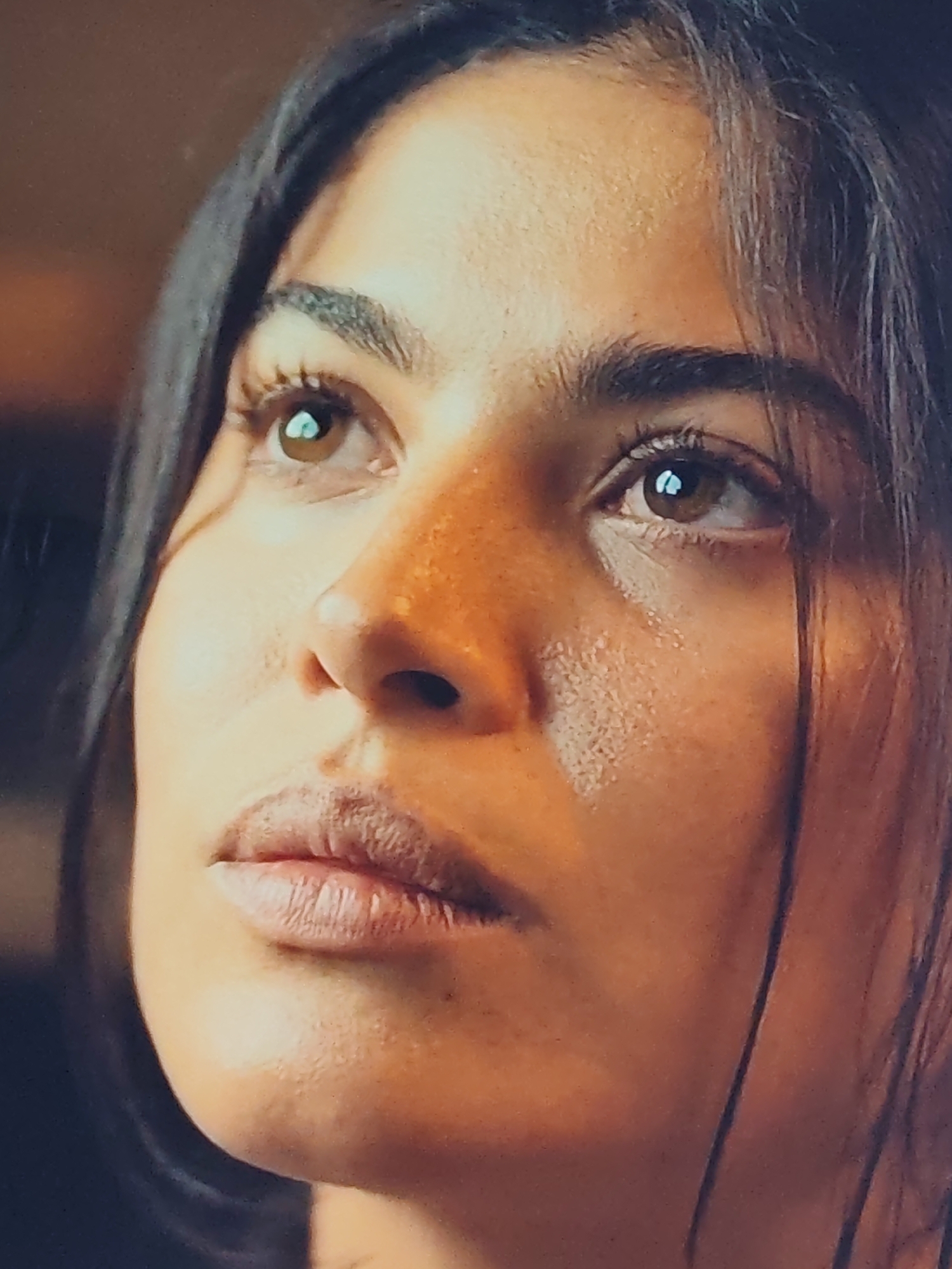 The image is a close-up, low-angle shot of a young woman's face, framed from the nose upwards. She has dark hair that frames her face and is looking upwards and to her left, with her eyes open. Her skin is warm-toned with visible pores and a slight sheen, suggesting natural light or possibly sweat. Her eyebrows are dark and well-defined, and her eyelashes are long and dark. Her eyes are a warm brown color, reflecting light. Her nose is prominent and slightly upturned. Her lips are full and appear slightly dry, with visible texture. The background is out of focus, appearing as a warm, diffused brown. The overall impression is one of introspection or observation. There is no indication of a specific city or location.