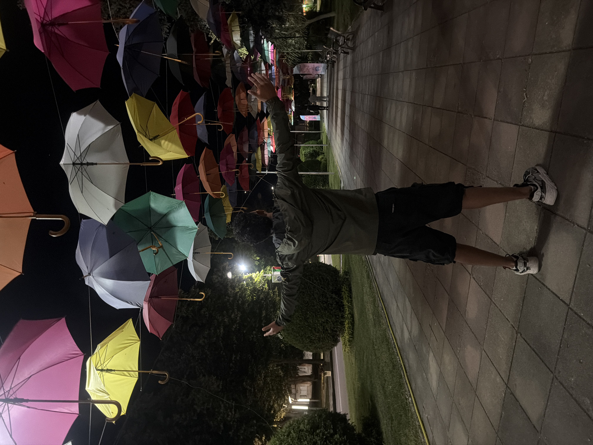 In Pavel Banya, Bulgaria, a young man stands with his arms outstretched, facing away from the camera, on a paved walkway lined with benches and lush green landscaping. Above him, a vibrant canopy of numerous colorful umbrellas is suspended, creating a festive and whimsical atmosphere. The scene is illuminated by ambient light, casting shadows on the walkway and highlighting the textures of the paving stones. The background reveals trees and buildings, suggesting an urban park or public space. The man is wearing a dark jacket and shorts, with athletic shoes on his feet. His pose implies a moment of joy or wonder, possibly interacting with the unique umbrella installation. The night sky is visible above the umbrellas, appearing dark.