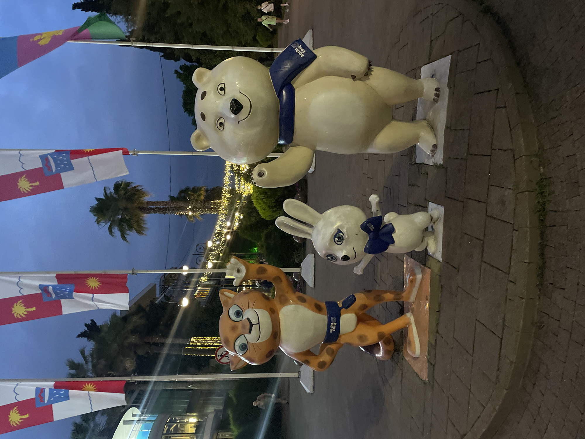 In Sochi, Russia, three large, cartoon-style statues of mascots stand on a paved walkway at dusk. From left to right, the statues depict a cheetah, a rabbit, and a polar bear. The cheetah is orange with black spots and wears a blue sash. The rabbit is white and wears a blue bow tie. The polar bear is white and wears a blue scarf. All three mascots are waving. Behind the statues, flags are displayed on poles, with palm trees and decorative lights visible. A building with lights is on the left, and trees line the background. A few pedestrians are visible in the distance. The sky is a deep blue, indicating it is evening.