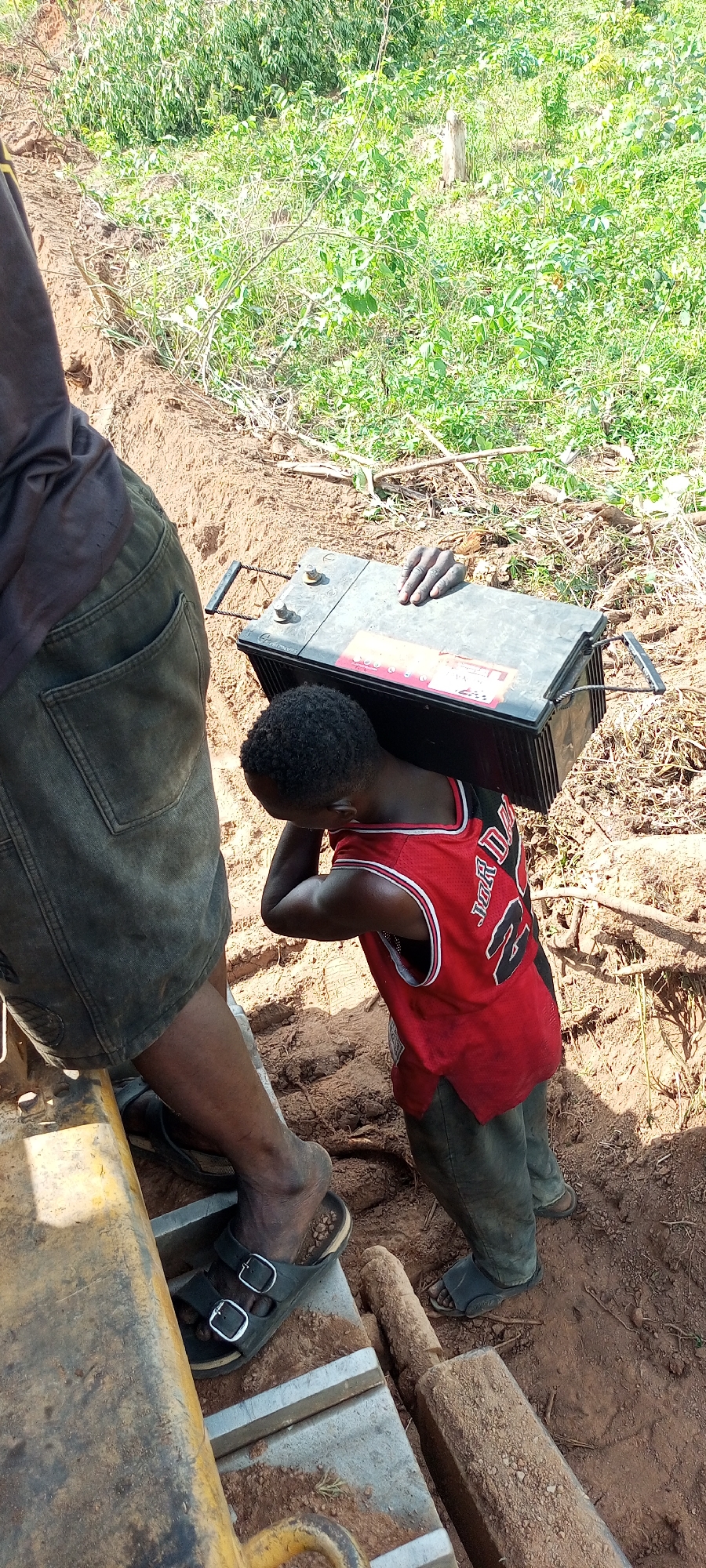 The image depicts a scene of manual labor in an outdoor, unpaved environment, likely during daytime given the bright natural light. The primary subject is a young child, possibly a boy, seen from the back and side. He is wearing a red sleeveless basketball jersey with "JORDAN" and the number "23" clearly printed in white on the back. The child is carrying a large, heavy-looking black battery on his head and right shoulder, steadying it with both hands. The battery features visible terminals and wire handles, and a red label with white, indistinct text is attached to its side.
Partially visible on the left side of the frame is an adult, whose legs and lower torso are shown. The adult is wearing dark shorts or trousers and black sandals with buckles, and is standing near a large, yellow piece of equipment, possibly machinery, which is also covered in dirt.
The ground is reddish-brown dirt, with a distinct trench or ditch running from the foreground towards the background. Beyond the immediate area of activity, lush green foliage and bushes are visible, indicating a rural or undeveloped setting. A wooden post or stake lies on the dirt near the adult's feet. The context provided indicates the location is Otunade, Nigeria.