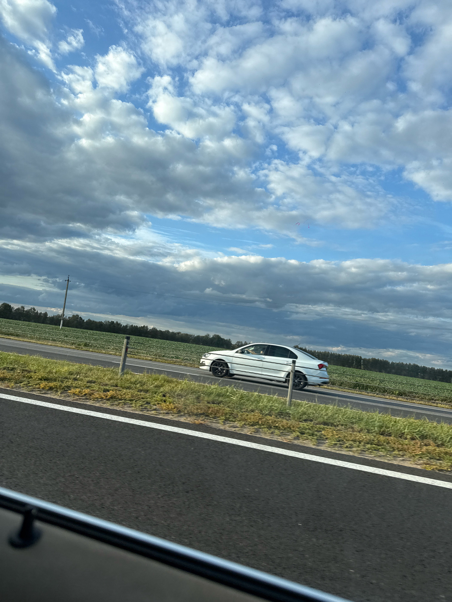 The image depicts a white sedan driving on a highway. The car is in motion, seen from the side, with its front wheels slightly turned. The highway has a paved surface and a white line marking the lane. Adjacent to the highway is a grassy verge and then a field of crops. In the distance, a line of trees borders the field, and a utility pole with wires is visible. The sky above is partly cloudy, with a mix of blue sky and white and grey clouds. The overall scene suggests a daytime, outdoor environment, likely a rural or semi-rural area, with the car traveling along a road.