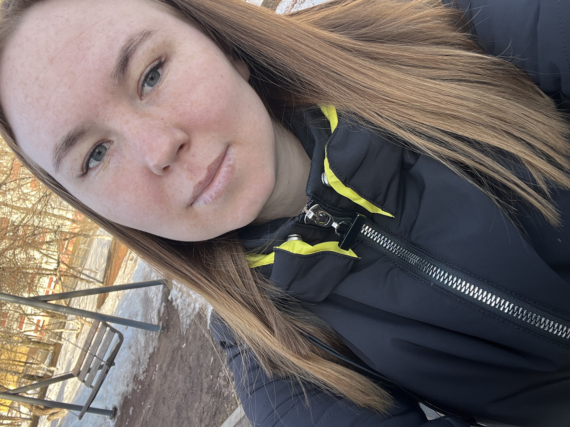A fair-skinned woman with light brown hair is facing the camera in an outdoor setting. She has freckles and is wearing a black puffer jacket with yellow accents. The jacket is partially zipped.
Behind the woman is a park-like area with a swing set visible to the left. There is melting snow on the ground, suggesting a cold season. Bare trees are visible in the background along with buildings.
Based on the description of the location being in the city of Chaikovsky, Russia, this scene likely depicts a park or recreational area in that city.