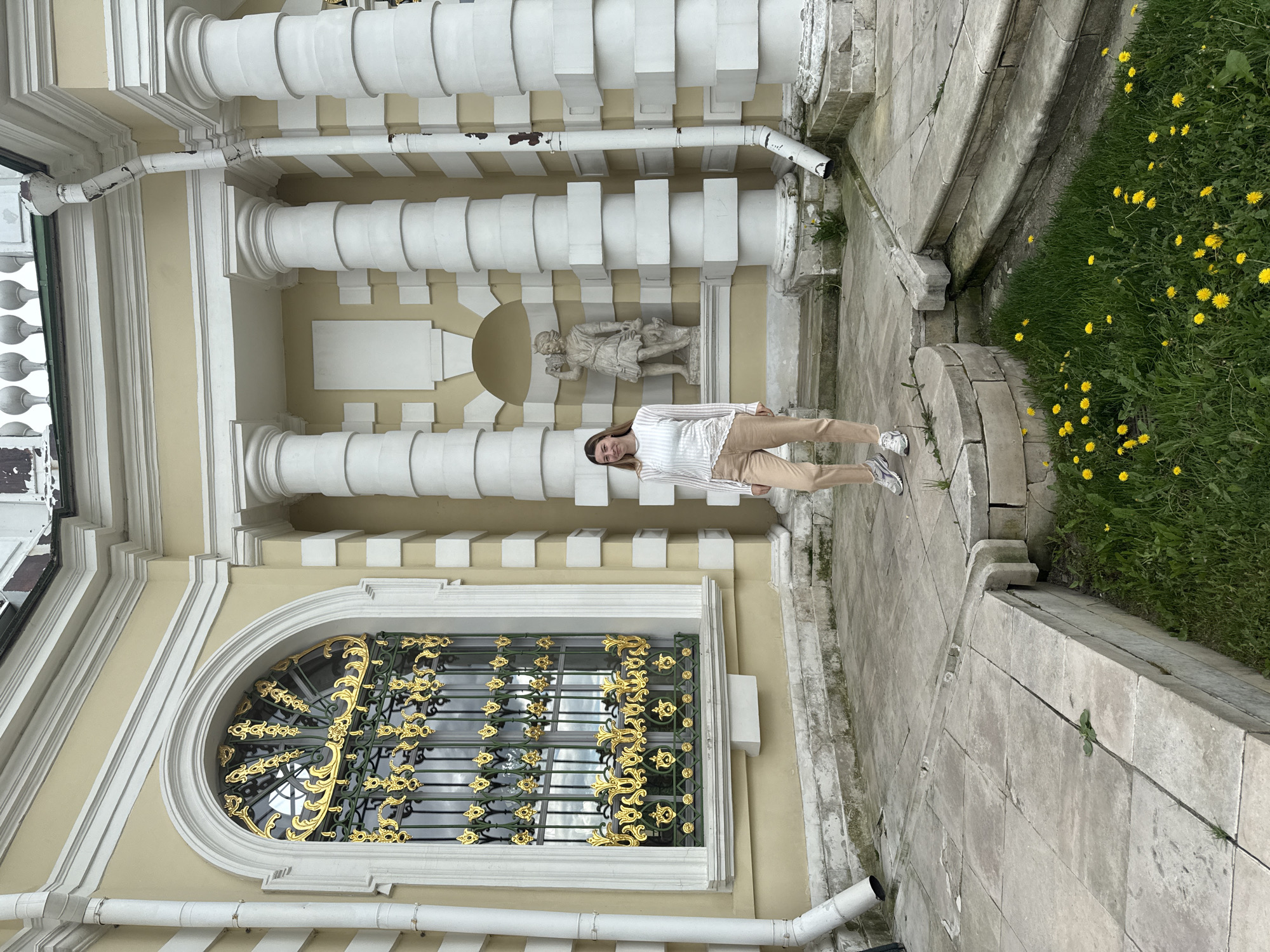 A young woman stands in front of the Ostankino Palace Museum in Moscow, Russia. The museum's facade features baroque architecture, including columns, a relief sculpture, and a window with ornate gold detailing. The surrounding area includes stone paving, steps, and a small patch of green with yellow flowers.