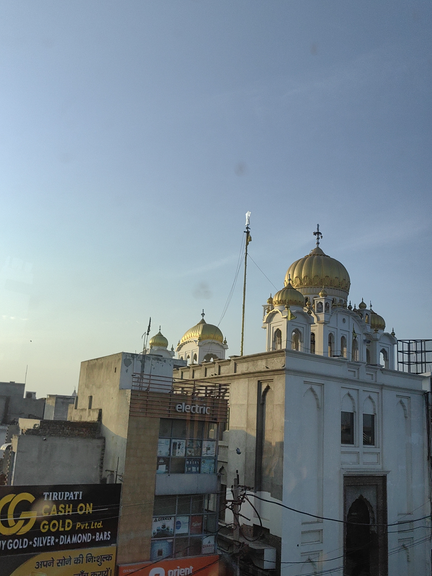 This image captures a view of the Gurudwara Shri Panipat Sahib in Panipat, India. The Gurudwara, a prominent Sikh religious site, is characterized by its distinctive golden domes and white architecture. In the foreground, the tops of various buildings are visible, including a multi-story structure with signage. One prominent sign reads "TIRUPATI CASH ON GOLD Pvt. Ltd." and advertises "MY GOLD * SILVER * DIAMOND * BARS". Another sign is partially visible with the word "orient". The sky above is a clear, pale blue, suggesting it is either early morning or late afternoon. The overall scene depicts a blend of religious architecture and urban commercial activity.