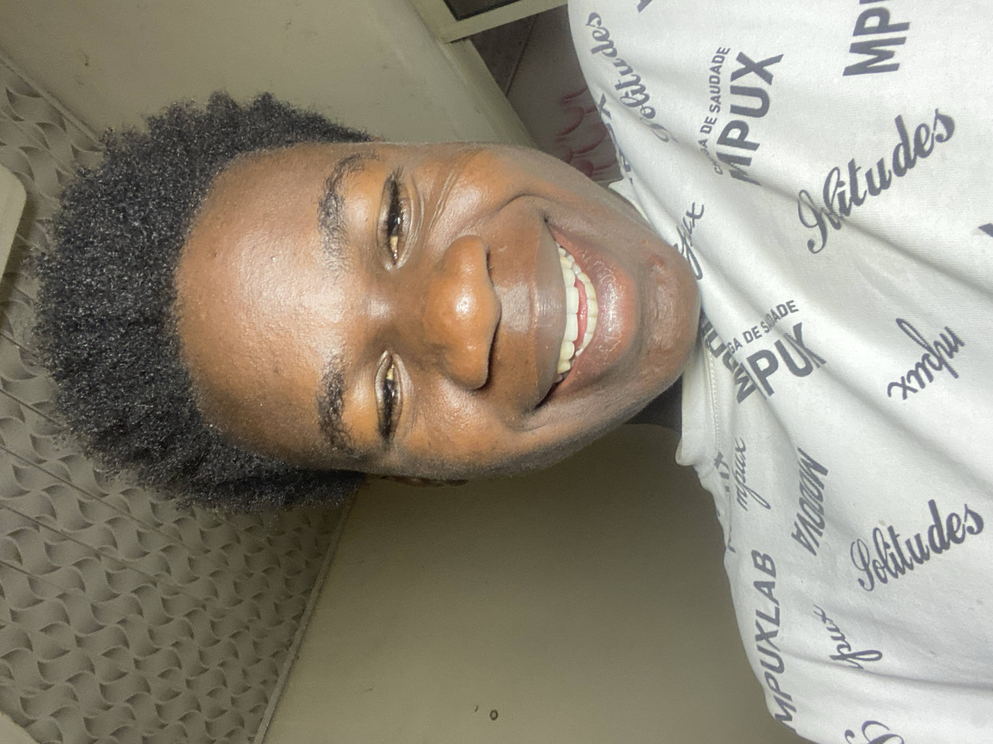 The image shows a close-up of a young Black man's face, smiling. He has short, dark, curly hair and is wearing a white shirt with the text "MPUXLAB" and "Solitudes" printed on it. The background appears to be an interior wall with a patterned texture. There is no location specified in the image but a city, Mararaba, Nigeria, is mentioned. Given the image content, there is no confirmation that the image originates from there.