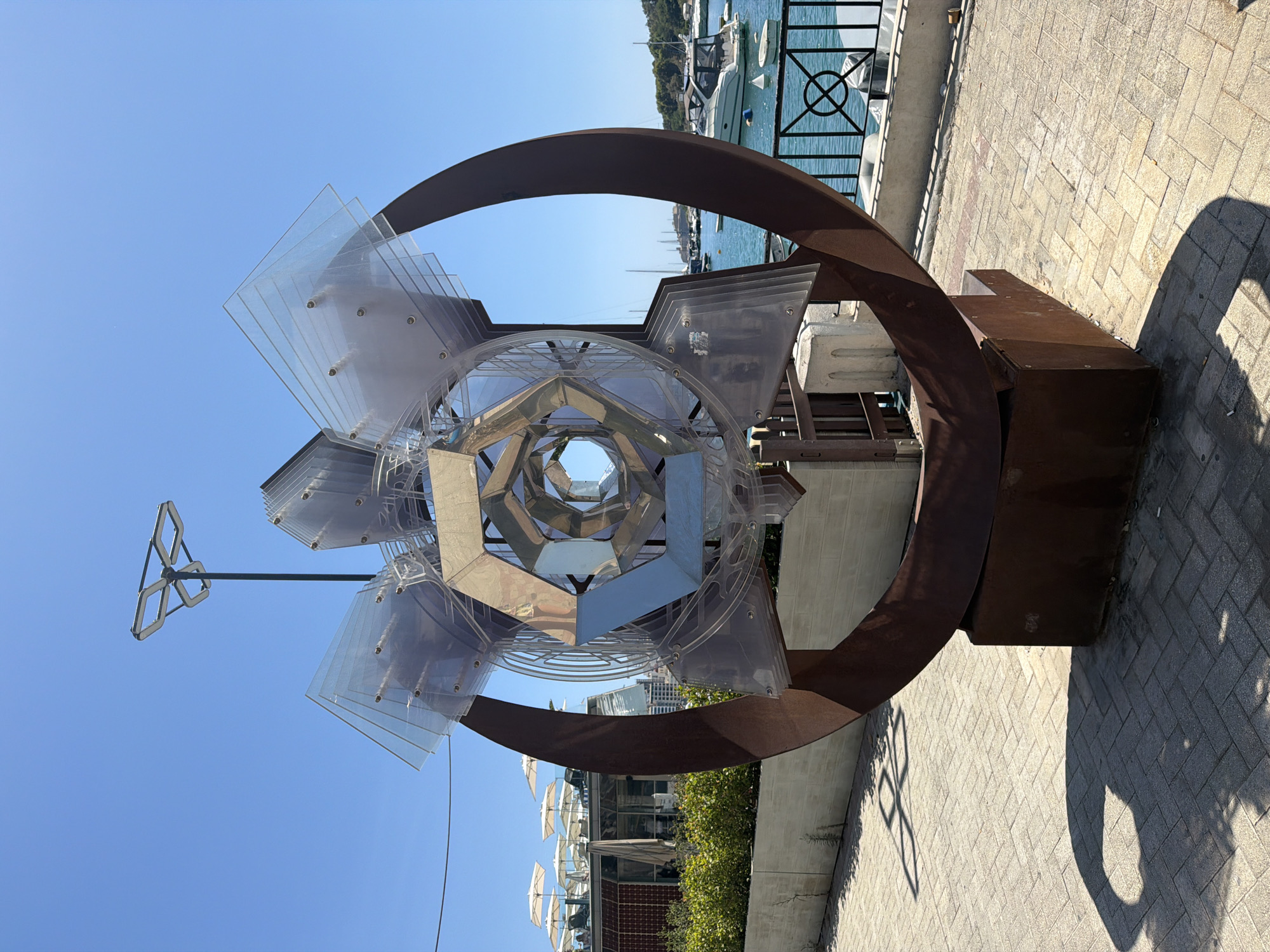 The image shows a large, abstract sculpture with a rusted metal base and a circular frame. Inside the frame is a complex structure made of transparent plastic and mirrored geometric shapes. The sculpture is located on a paved walkway next to a body of water, likely the Sliema Creek in Gżira, Malta, as suggested by the presence of boats and the distinctive waterfront architecture in the background. The sculpture casts a long shadow on the ground, indicating it is a sunny day. In the background, there is a building with a rooftop terrace occupied by people under umbrellas. The water is calm and several boats are docked or anchored. The sky is clear and blue.