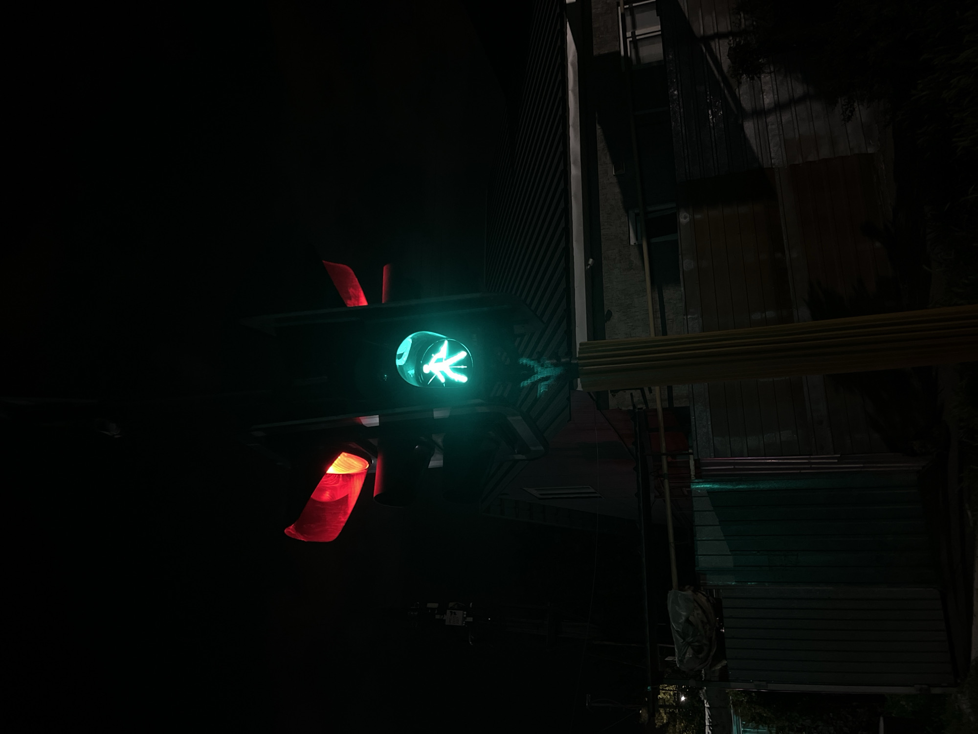 This image depicts a nighttime street scene in Almaty, Kazakhstan, centered on a set of illuminated traffic signals. On the left, a vehicular traffic light assembly displays two active red signal lights, indicating a stop command for vehicles. To the right of the vehicular traffic light, a pedestrian signal is illuminated green, displaying the iconic "walking man" symbol, which permits pedestrian crossing. The background is enveloped in darkness, characteristic of nighttime. A building, likely a residential structure, is visible in the mid-ground, featuring a dark, horizontally ribbed roof and a single illuminated window on its side. Below this building, various fences and structural elements are present, including a section of corrugated metal fencing. Utility poles and overhead wires are discernible on the far left side of the frame. Sparse, dark vegetation is visible in the lower portions of the image. No individuals are present, and no specific actions or interactions are depicted.