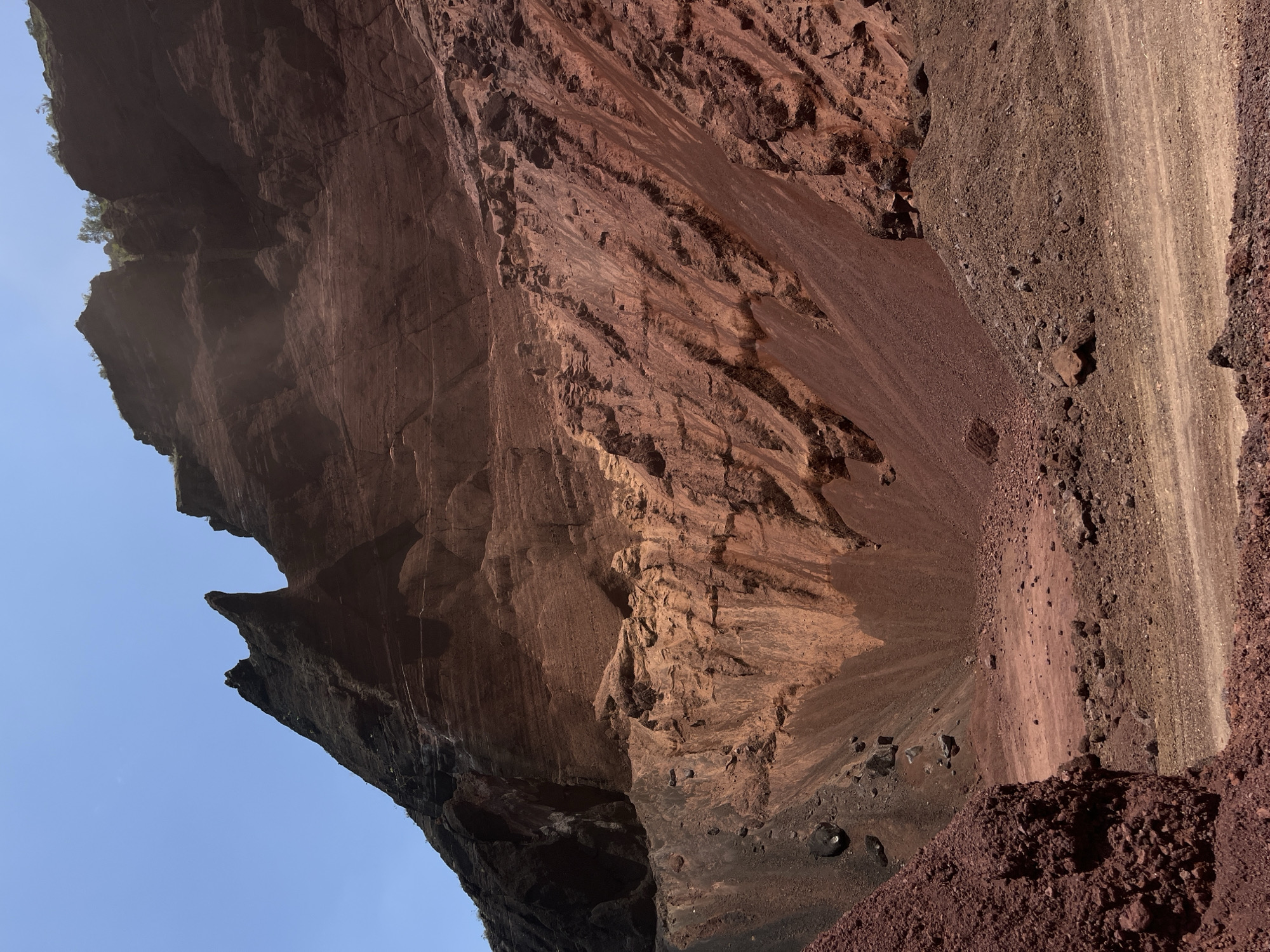 The image shows a volcanic crater with steep, eroded walls displaying layers of red and black rock. The ground within the crater is composed of loose gravel and dirt, with scattered rocks. A clear blue sky is visible above the rim of the crater. The scene depicts a natural landscape devoid of any human presence or activity, characteristic of a geological formation in San Miguel Papaxtla, Mexico.
