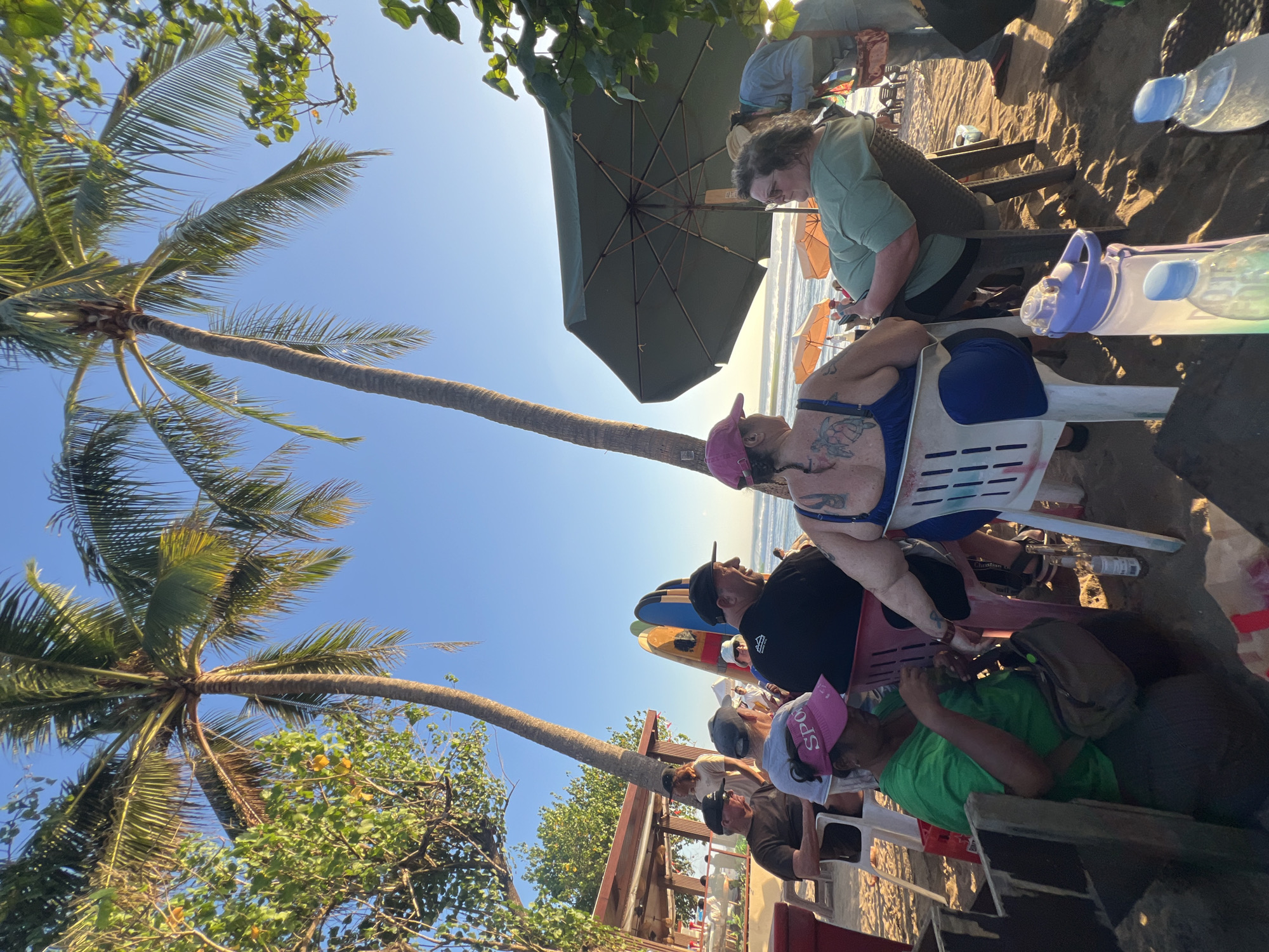 This image depicts a beach scene in Kuta, Indonesia, likely during the late afternoon or early evening, as indicated by the low angle of the sun and the warm lighting. Several people are seated on chairs on the sand, facing the ocean and the setting sun. Palm trees are prominent in the foreground and background, with their fronds silhouetted against the clear blue sky.
In the mid-ground, a group of people are gathered, some appearing to be socializing. Several surfboards are leaning against a structure behind them, suggesting a popular surf spot. An open-air establishment with a roof and tables is visible to the left, possibly a beachside cafe or bar. Beach umbrellas are scattered on the sand in the distance, and the ocean waves are visible.
The people in the foreground are diverse in age and attire. One person is wearing a green t-shirt and a pink visor, holding a bottle. Another individual, seated to the right, is wearing a green t-shirt and appears to be looking towards the ocean. A person in a dark shirt and black baseball cap is seated in the front left, looking to their right. Another person in a black shirt and black baseball cap is seated in the front right, looking towards the ocean. A woman with a large tattoo on her back is seated in a white chair, wearing a blue bathing suit. In the background, several other individuals are visible, some standing and some seated, and the general atmosphere seems relaxed and social. The presence of water bottles and other personal items indicates a leisurely outing.