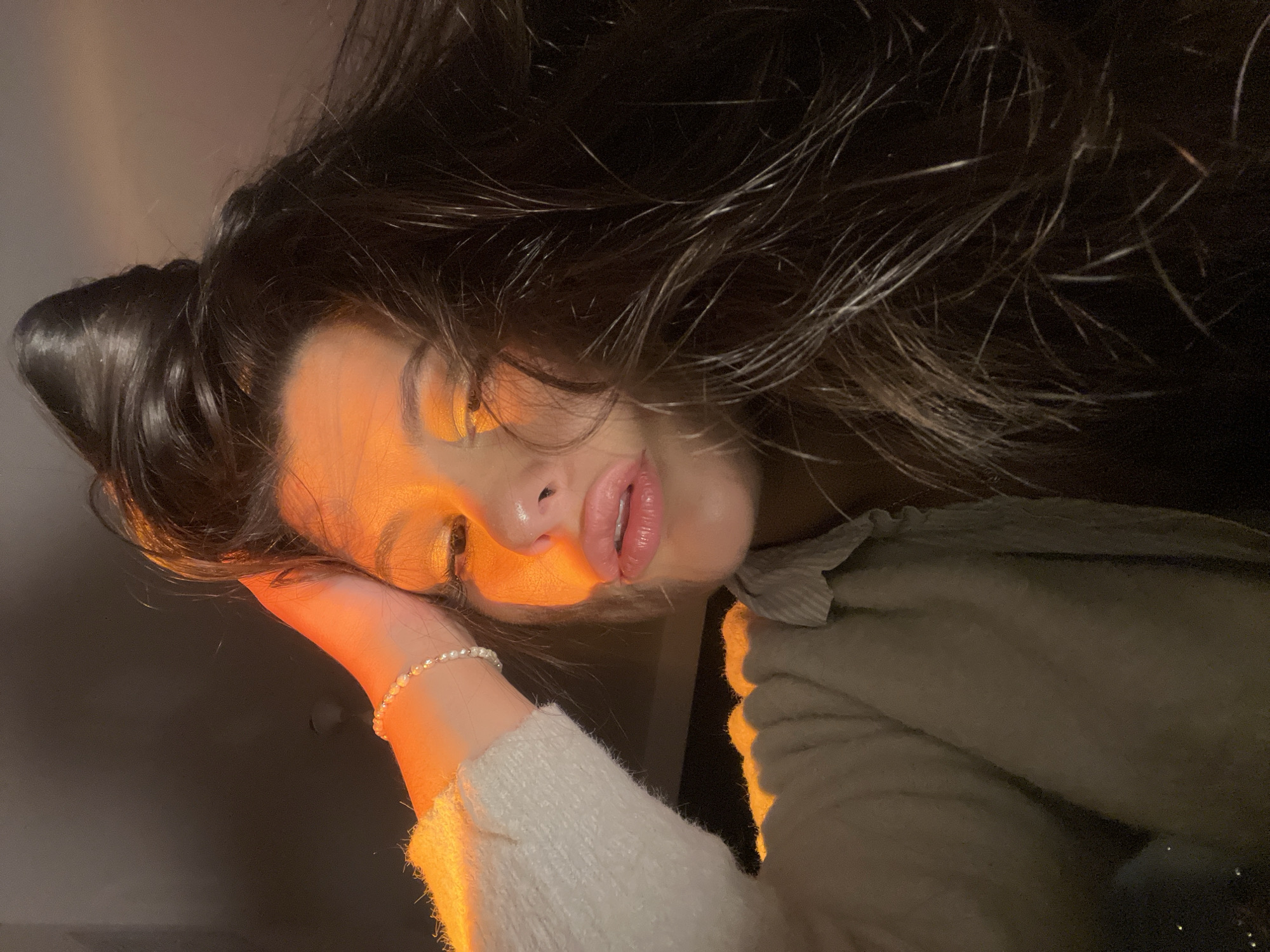 The image depicts a close-up, angled portrait of a single female-presenting individual, illuminated primarily by a strong orange-amber light source on the left side of the face and arm. The subject has dark, long hair, styled with volume on top, and appears to be looking towards the camera. Her lips are full, slightly parted, and coated with a glossy substance. Her left eye is partially visible, featuring what appears to be orange or gold eyeshadow. The left hand, adorned with a pearl bracelet, is raised towards the head, with fingers partially obscured by hair. She is wearing a light-colored, possibly cream-toned, textured long-sleeved garment, with a lighter, possibly striped, shirt visible underneath. The background is indistinct and dark, indicating an indoor or low-light environment. The scene is situated in Kiryat Bialik, Israel.