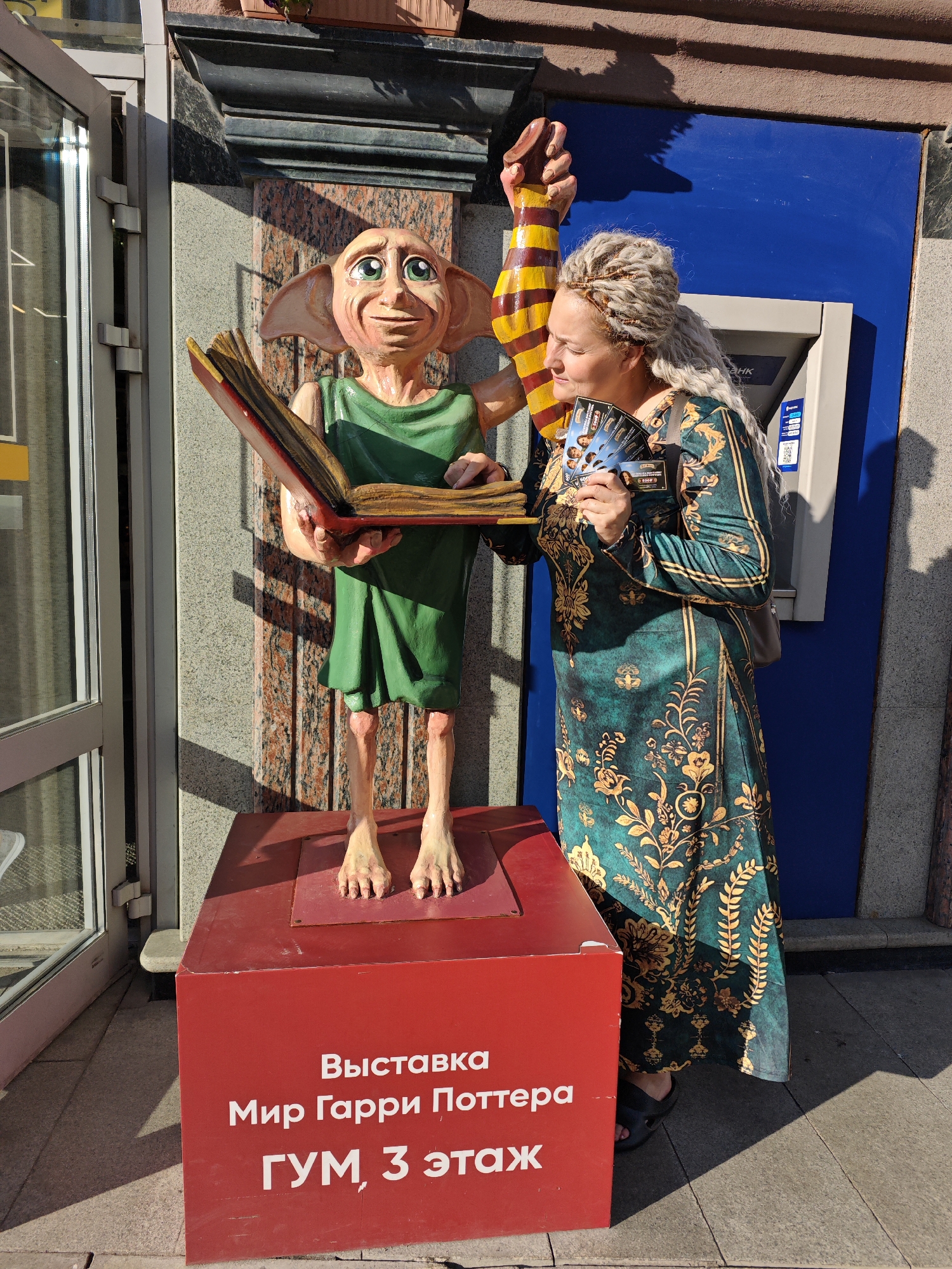 A woman with blonde braided hair and a long, patterned green dress stands next to a statue of Dobby from Harry Potter. Dobby is a light-skinned, elfin figure with large ears, wearing a green tunic and holding an open book. The woman is holding several small flyers in her right hand, and appears to be looking at them. She is standing on a paved sidewalk in front of a building with blue and grey facades. A red pedestal is in front of the statue, with white text on its side that reads "Выставка Мир Гарри Поттера ГУМ, 3 этаж" (Exhibition Harry Potter World GUM, 3rd floor). An ATM is visible on the blue facade of the building behind the woman. The scene suggests an outdoor exhibition or event related to Harry Potter, likely in a public space. The text on the pedestal indicates the location is GUM, on the 3rd floor, and it is an exhibition of the "World of Harry Potter." GUM is a well-known department store in Moscow, but this text is in Russian, and the style of the building and the presence of an ATM suggest a commercial area in a Russian city. A search for "Harry Potter exhibition GUM Kazan" confirms that such an exhibition took place at the GUM department store in Kazan, Russia.