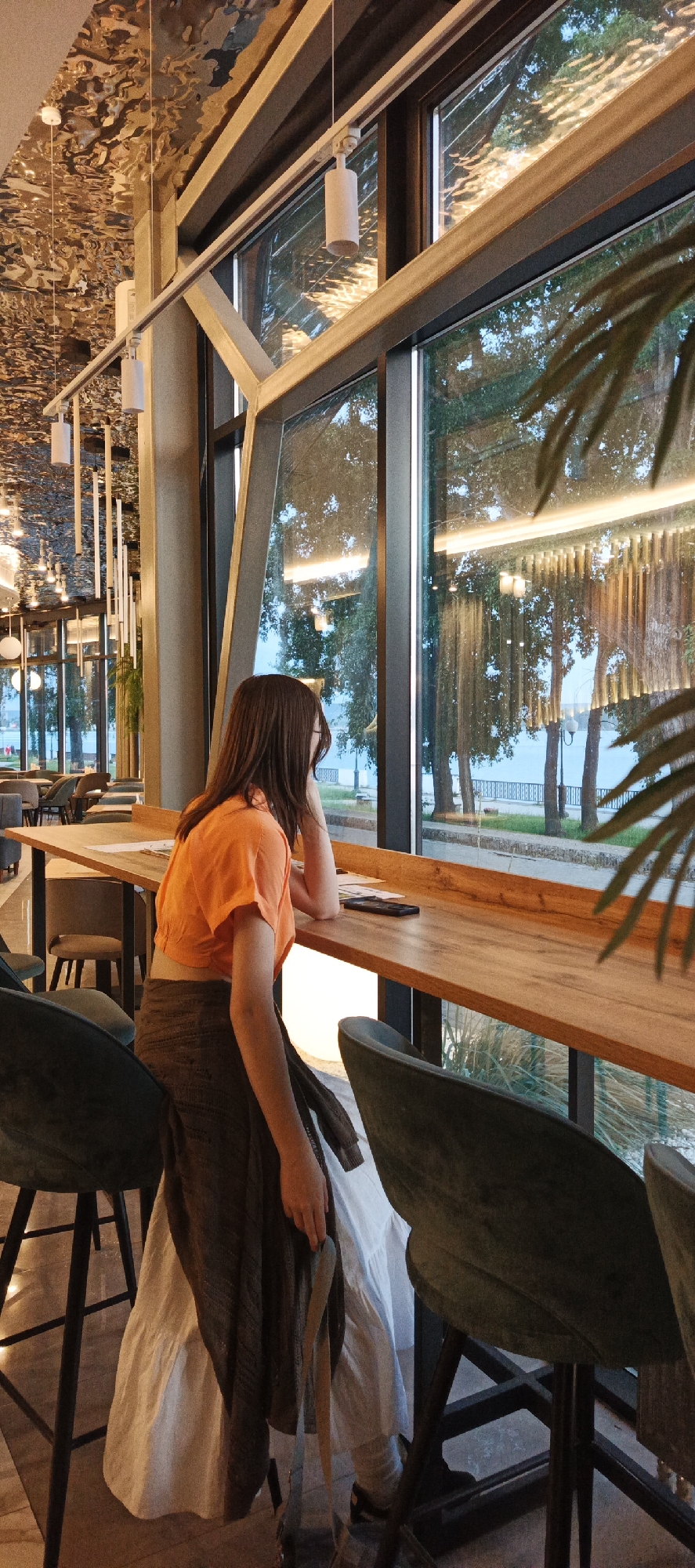 A young woman with long, dark hair is sitting at a counter in a cafe, looking out a large window at a body of water and trees. She is wearing an orange crop top and a long, flowing white skirt, with a dark brown cardigan draped over her shoulders. The cafe appears to be modern, with a decorative ceiling and industrial-style lighting. The window offers a view of Votkinsk, Russia, with its characteristic waterfront scenery. The overall atmosphere suggests a moment of quiet contemplation.