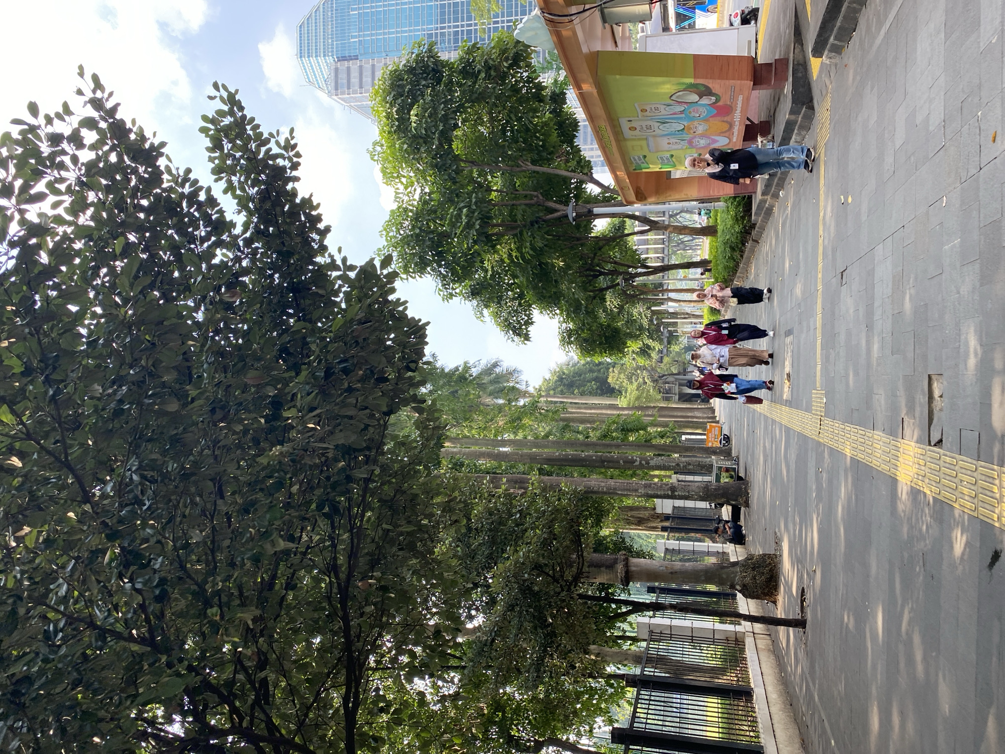 The image depicts a sidewalk scene in the city of South Jakarta, Indonesia. A group of five women wearing hijabs walks along the sidewalk. To the right of the sidewalk is a bus stop shelter with a colorful advertisement featuring what appear to be cosmetic products. A woman in a black shirt and blue jeans stands near the bus stop, looking towards the camera and holding her hand to her face. In the background, a tall, modern glass skyscraper rises into the sky, suggesting an urban setting. Trees line the sidewalk, casting shadows on the pavement. A black fence runs along the left side of the sidewalk, and beyond it, more trees and vegetation are visible. A person is seated on the ground near a tree to the left, appearing to be using a mobile phone. The ground is paved with grey tiles and features a yellow tactile paving strip for visually impaired individuals. The sky is partially cloudy.