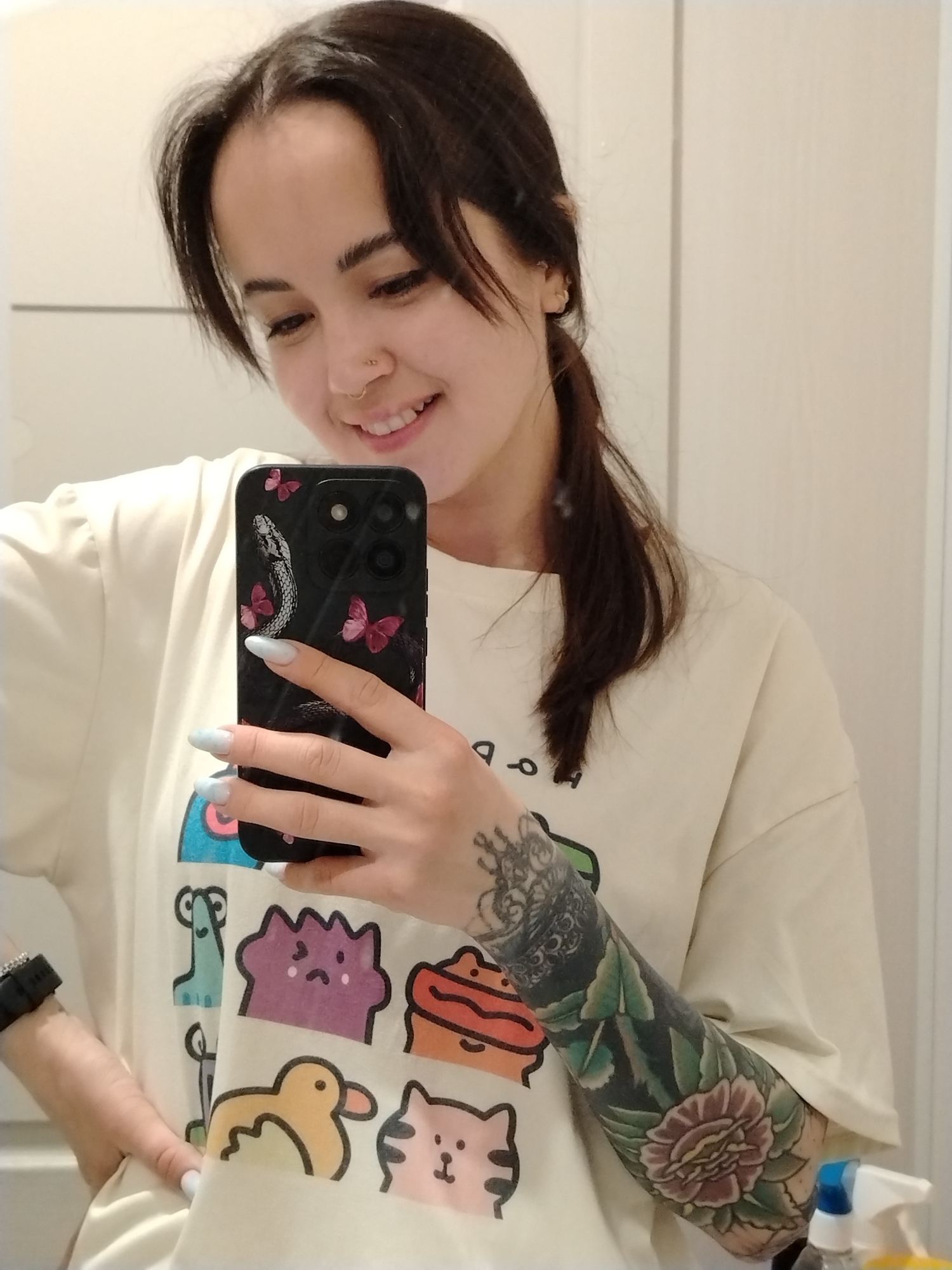 A young woman is captured in a mirror selfie, smiling softly while holding a black smartphone. She has dark hair styled with crimped roots and a ponytail, and a small stud nose piercing. She wears a cream-colored, short-sleeved t-shirt adorned with whimsical, colorful cartoon characters, including a blue monster, a purple blob, an orange creature, a yellow duck, and a pink cat. Her nails are painted a light periwinkle blue.
Her left arm is heavily tattooed with a full sleeve featuring intricate floral designs, predominantly a large rose in shades of red and pink with green leaves, and an ornate crown-like pattern on the upper arm. A black digital watch or fitness tracker is visible on her left wrist. The smartphone she holds has a black case with a distinct design of a snake coiling around it, interspersed with several pink butterflies. She is looking down towards the phone, her expression gentle and content. The setting appears to be indoors with plain light-colored walls, possibly reflecting surfaces, and a blue and white spray bottle is partially visible in the bottom right corner.