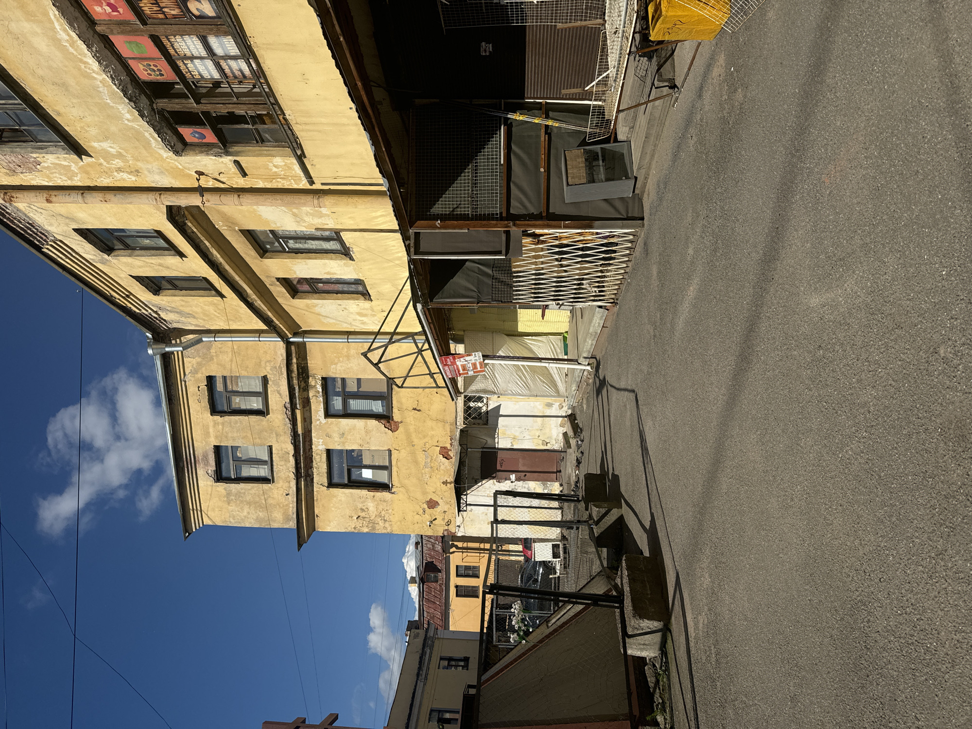 The image depicts a narrow urban courtyard in Saint Petersburg, Russia, bathed in sunlight. The dominant structure is a weathered, yellow apartment building with a peeling facade, featuring multiple windows, some of which are boarded up or covered with paper. A rusty downspout runs vertically along the building. In the foreground, a paved asphalt surface forms a walkway or courtyard. To the left, a chain-link fence encloses an area with a large, triangular, canvas-covered object and concrete blocks. Further back, more fencing is visible, along with a dark brown door on the yellow building. Adjacent to the yellow building, a covered walkway with open stalls and metal grating is present. A white accordion-style gate is positioned in front of one of these stalls. A metal stand with a wire shelf holds yellow objects, one of which appears to be a large plastic bin. The sky above is a vibrant blue with a few scattered white clouds. The overall scene suggests an older, somewhat neglected urban environment, possibly a back alley or service area of a historic building.