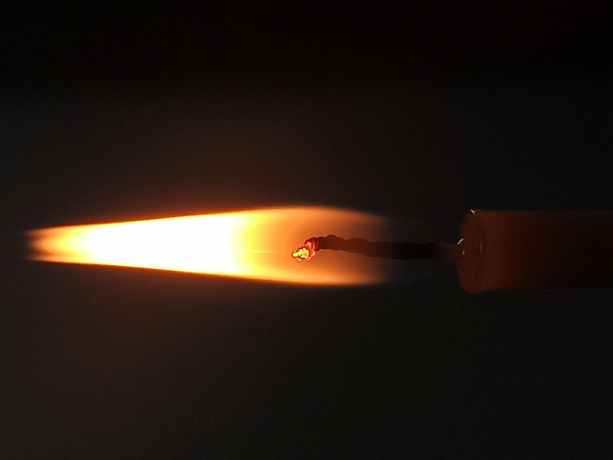 A close-up, vertical shot captures the vibrant flame of a single candle against a dark, blurred background. The flame, predominantly a bright yellow and white, tapers to a pointed tip. A small, glowing ember is visible at the base of the flame, where it meets the wick, which appears dark and slightly charred. Below the flame, the upper portion of the candle is visible, a soft orange hue indicating its waxy composition. The overall impression is one of warmth and focused light against a void. The location is Unknown, Unknown.