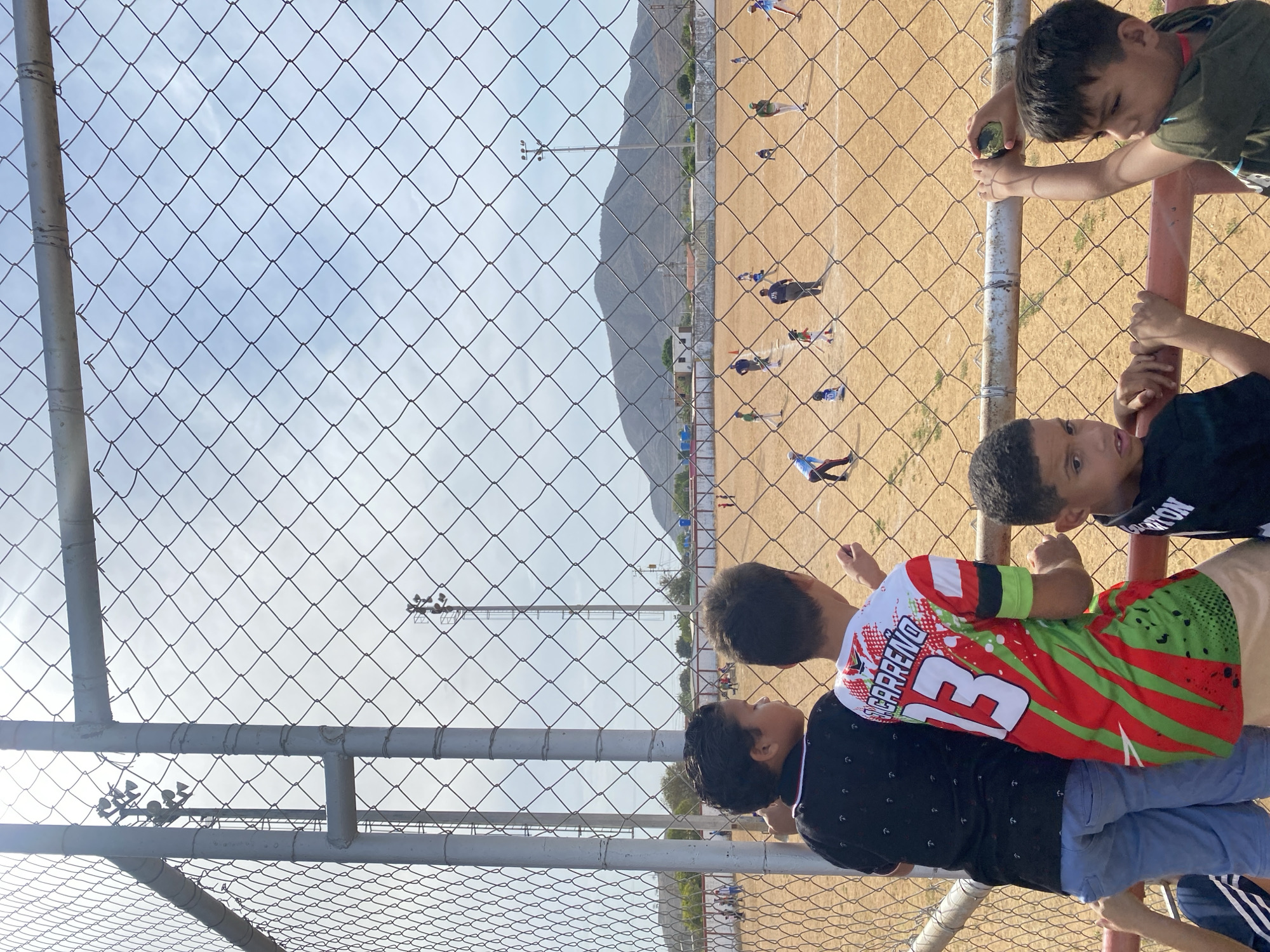 Several children stand behind a chain-link fence, observing a baseball field. The field is dry and dusty, with players in various positions. In the background, a mountain range is visible under a bright sky with some clouds. The location is near or within Juan Griego, Venezuela.