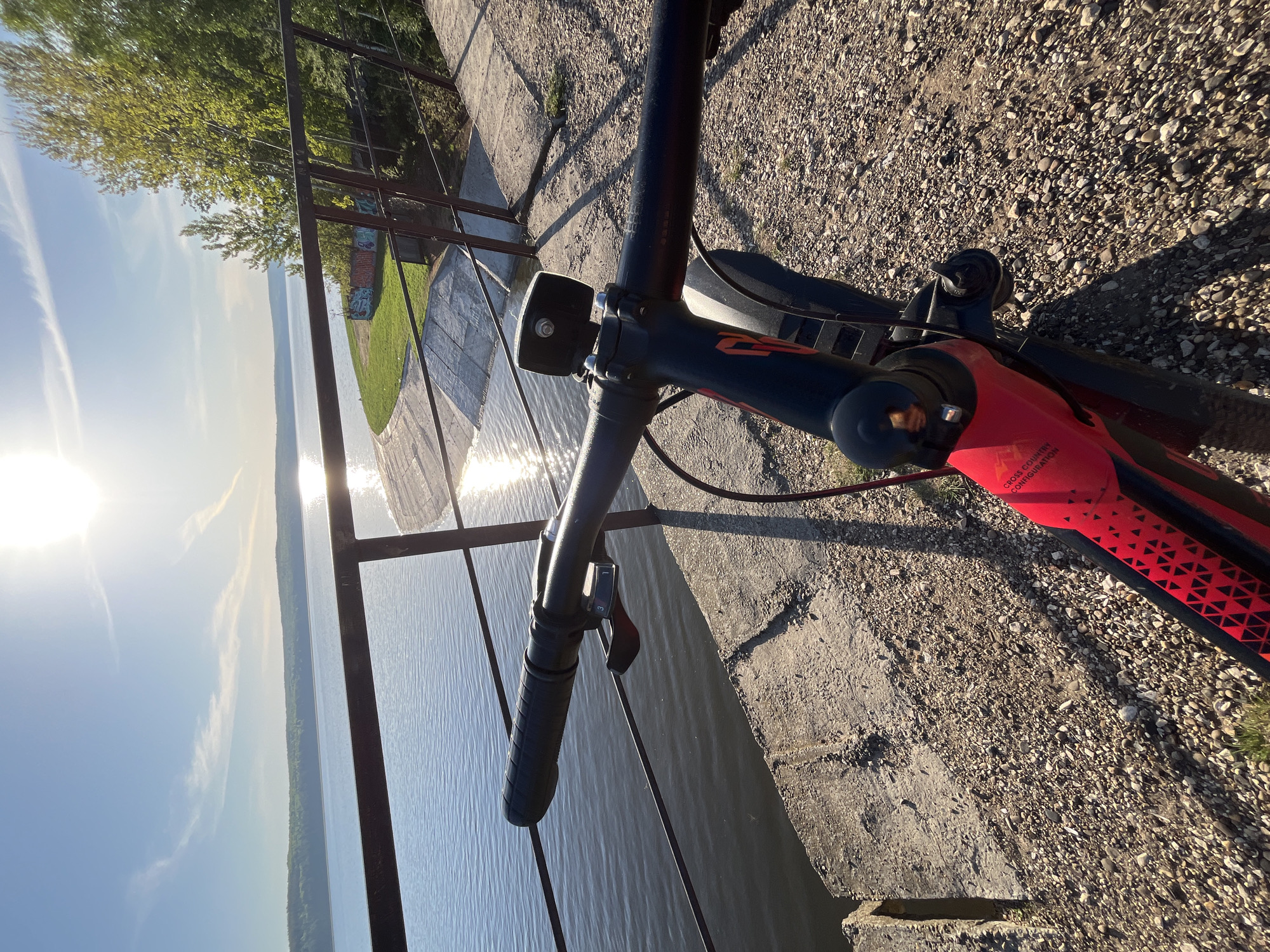 A bicycle is perched on a concrete ledge, with its handlebars and front portion dominating the frame. Beyond the bicycle, a metal railing separates the viewer from a body of water, reflecting the sunlight intensely. The horizon reveals a stretch of land with sparse vegetation and structures, hinting at the city of Chaikovsky, Russia. The foreground is composed of a gravel surface, contrasting the smoothness of the water and the concrete structures.