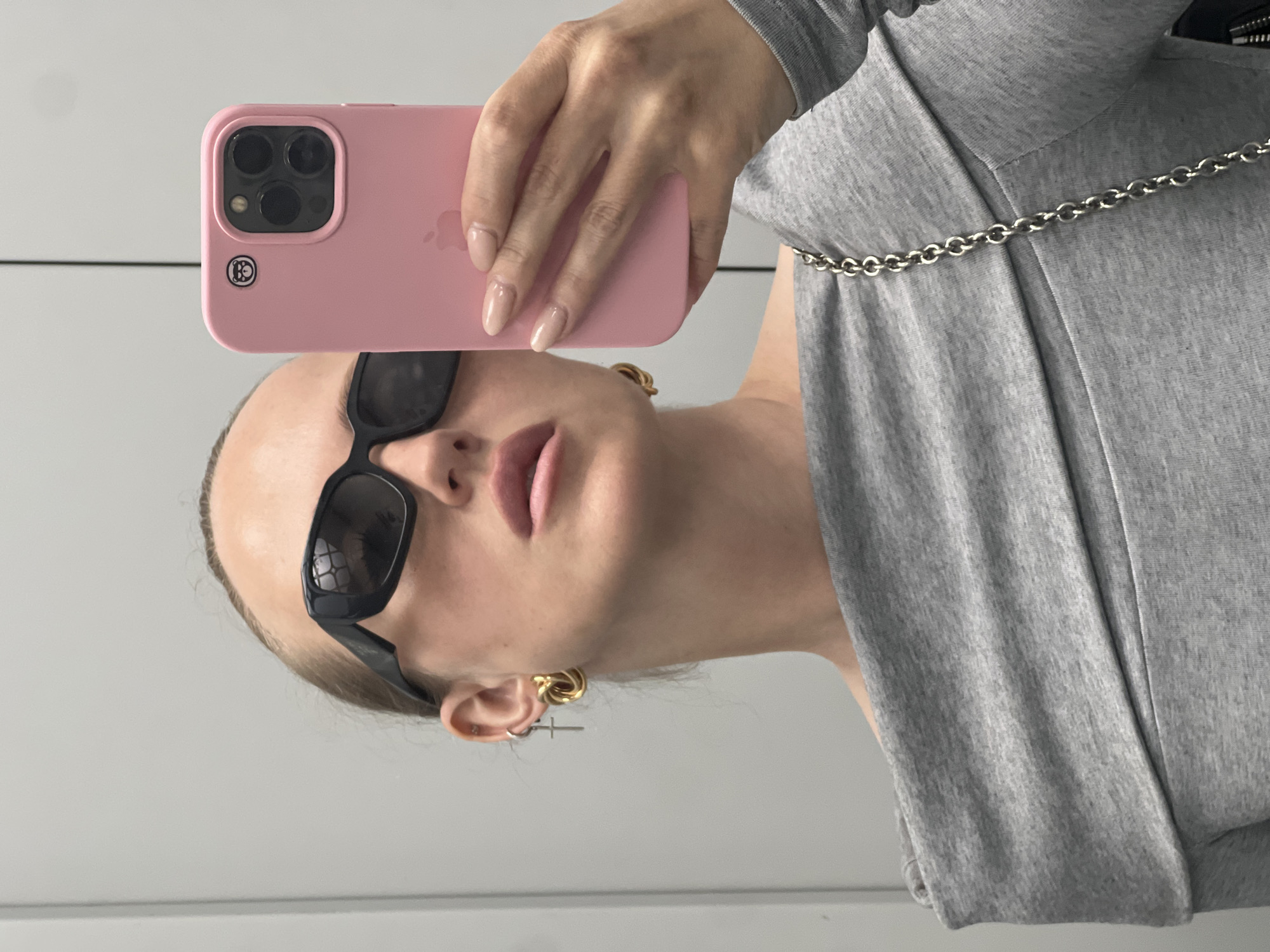 A woman is taking a selfie in a mirror. She is wearing black sunglasses, gold knot earrings, and a grey off-the-shoulder top with a silver chain strap. She is holding a pink iPhone in her right hand, showing the camera to the mirror. Her nails are painted a neutral color. The background is a plain, light-colored wall. The image is taken from the chest up.